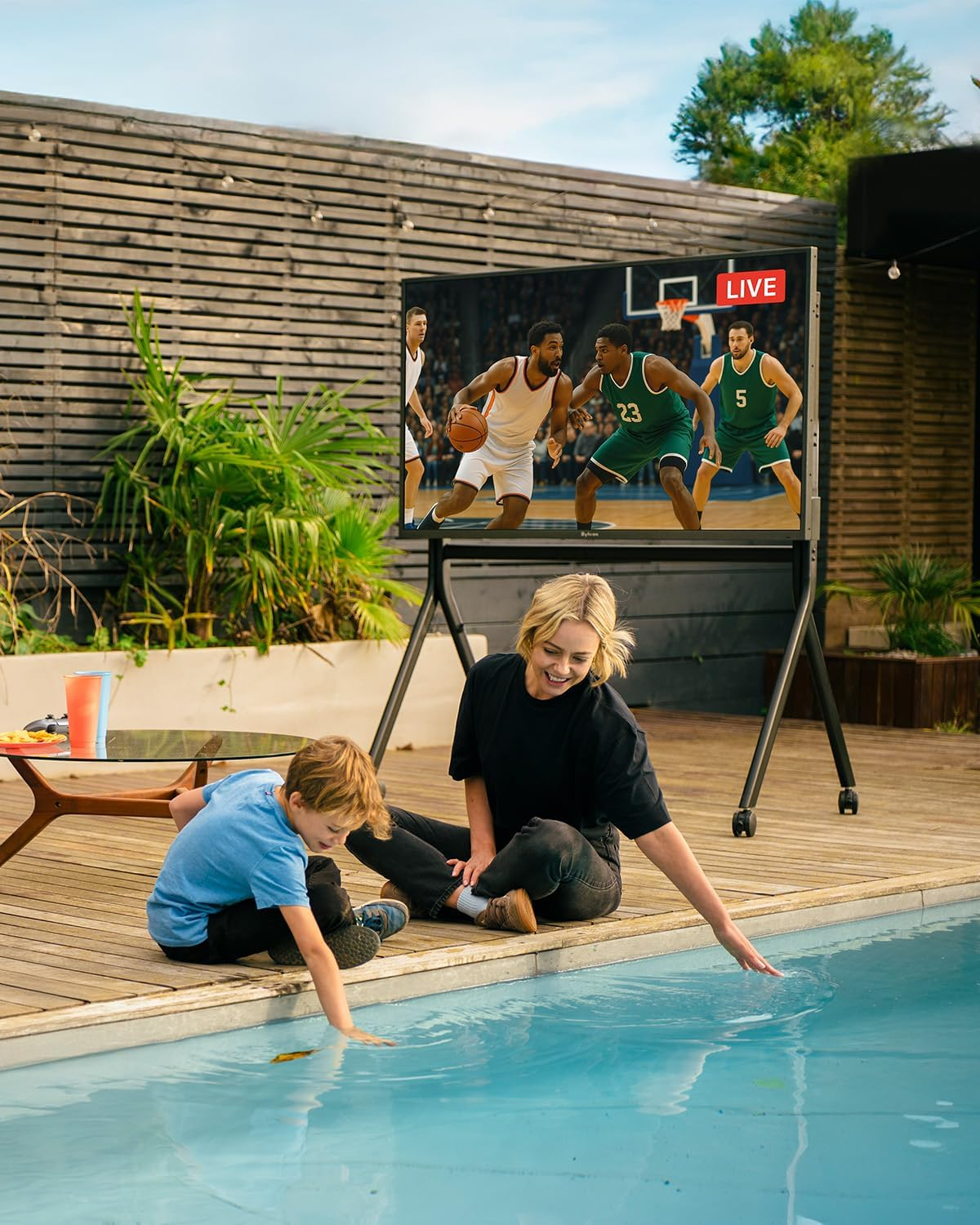 Una mujer y un niño junto a una piscina, con un televisor grande mostrando un partido de baloncesto.