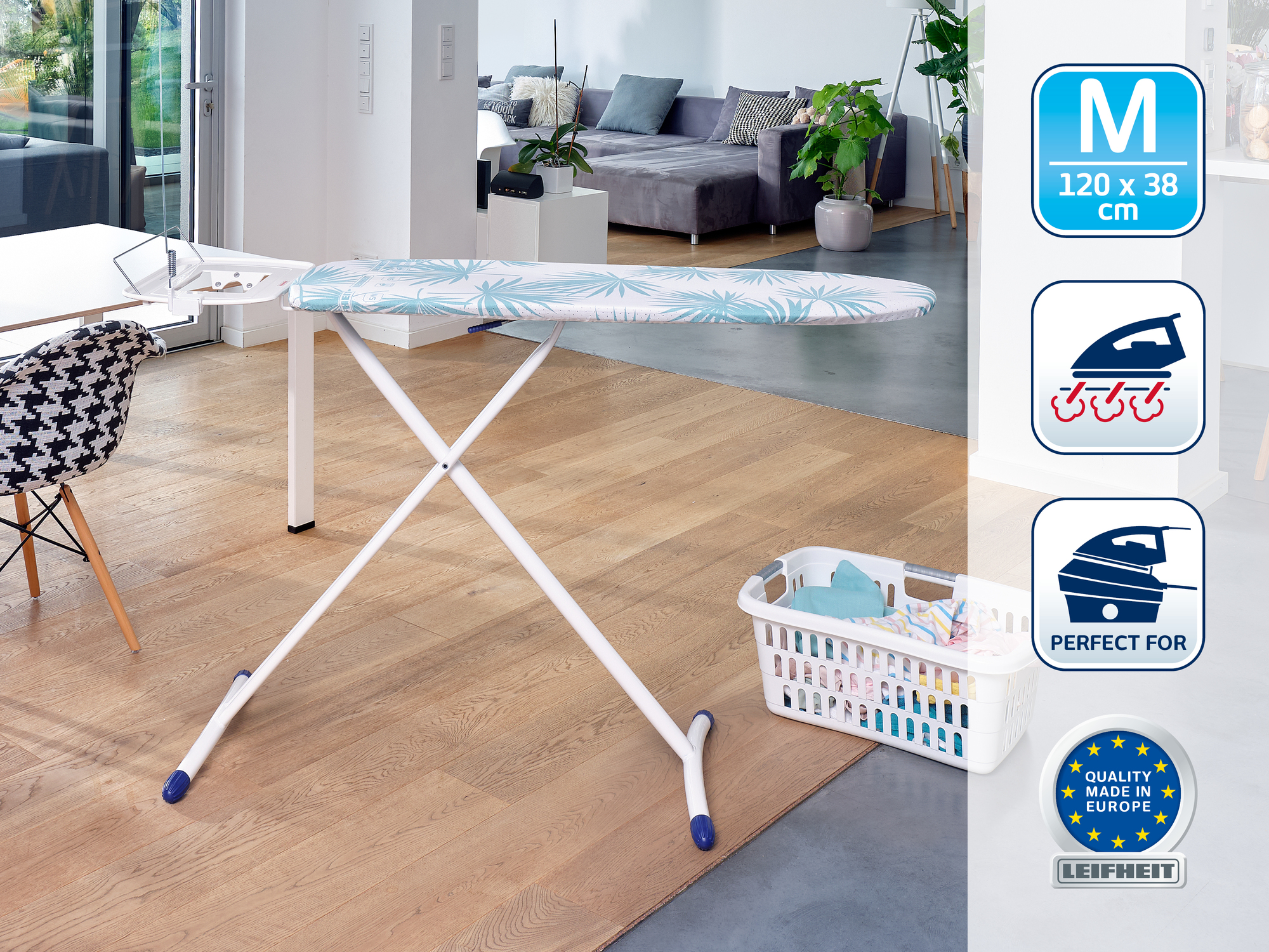 Una tabla de planchar blanca con patrón azul sobre suelo de madera en una sala, con una cesta de ropa al lado.
