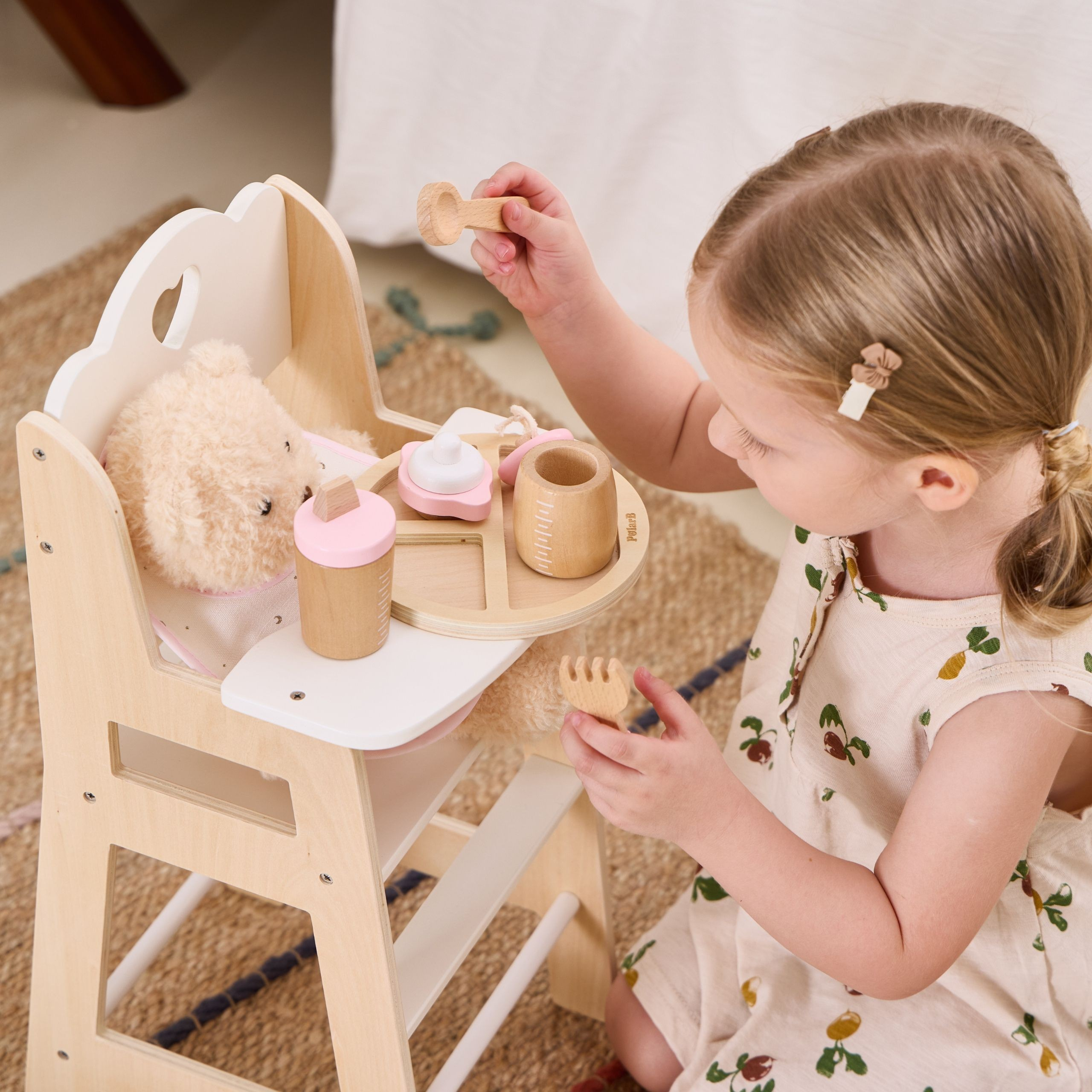Ein blondes Mädchen füttert einen Plüschbären in einem Holz-Hochstuhl mit Spielzeugutensilien.