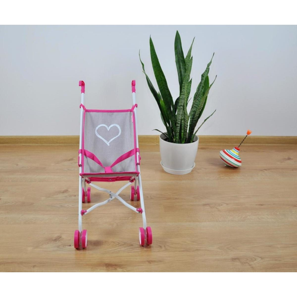 Ein pink-grauer Puppenbuggy mit Herzmuster auf Holzfußboden, daneben eine Topfpflanze und ein Kreisel.