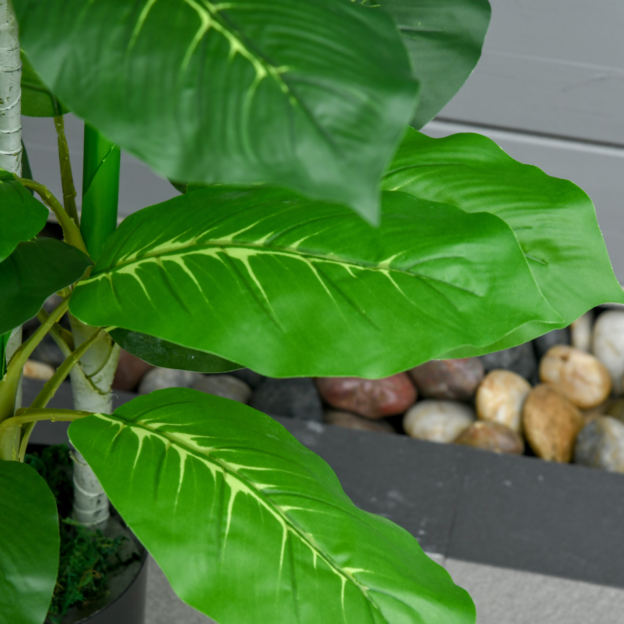 Primer plano de una planta verde artificial con hojas grandes y nervadas frente a un borde gris y guijarros.