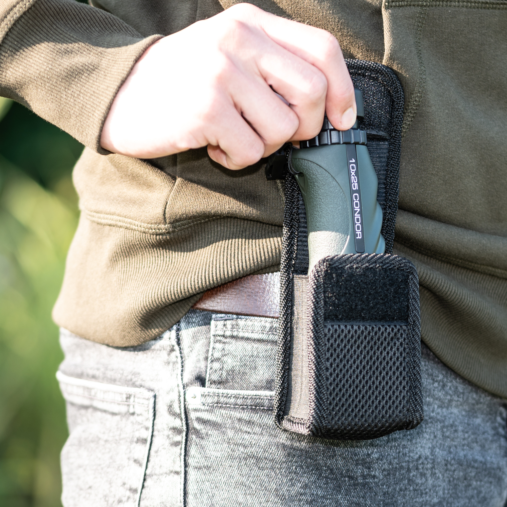 Una persona con chaqueta verde saca un monocular Condor 10x25 de una funda negra en su cinturón.