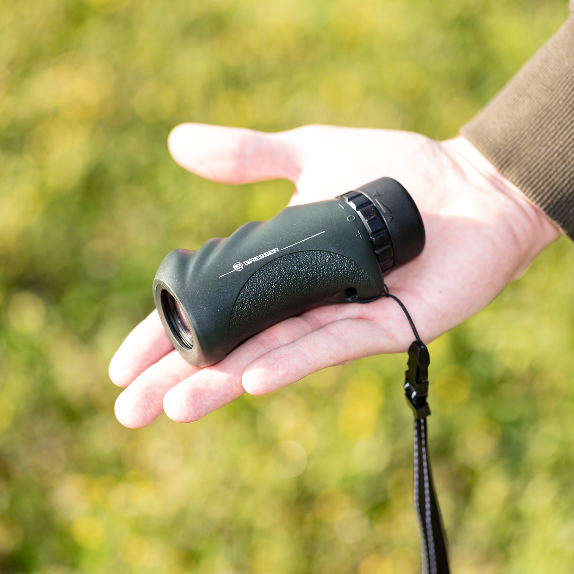 Una mano sostiene un monocular Bresser compacto y verde oscuro sobre un fondo verde borroso.