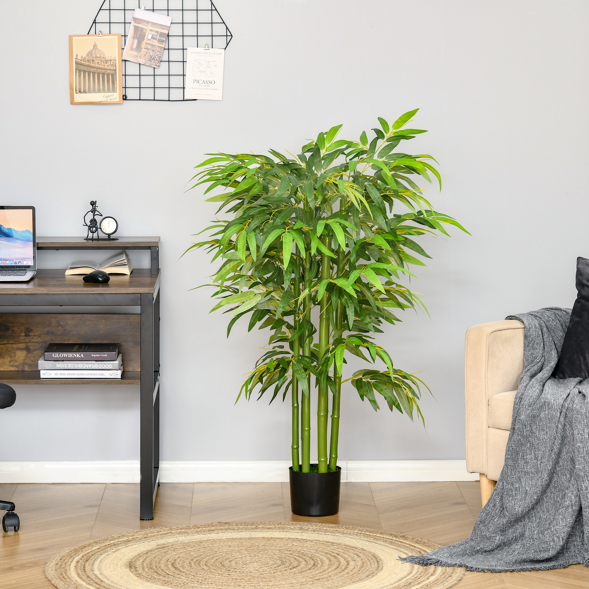 Una planta de bambú artificial verde en una maceta negra en el interior, junto a un escritorio y un sofá con manta.