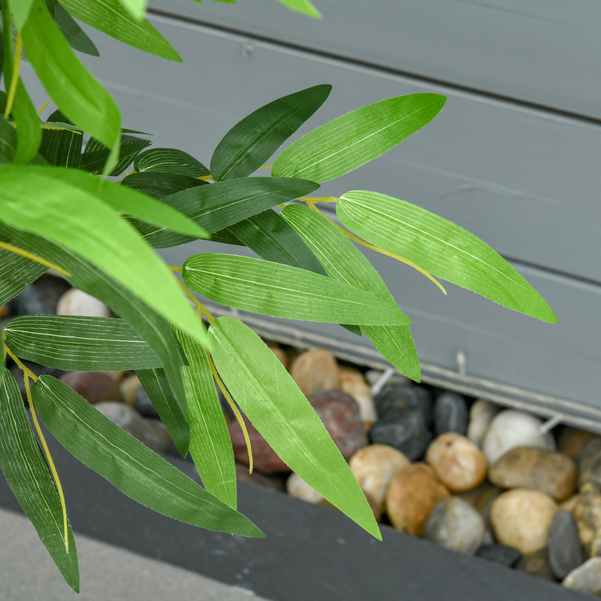 Hojas de bambú verdes en primer plano con una valla de madera gris y guijarros de río detrás.