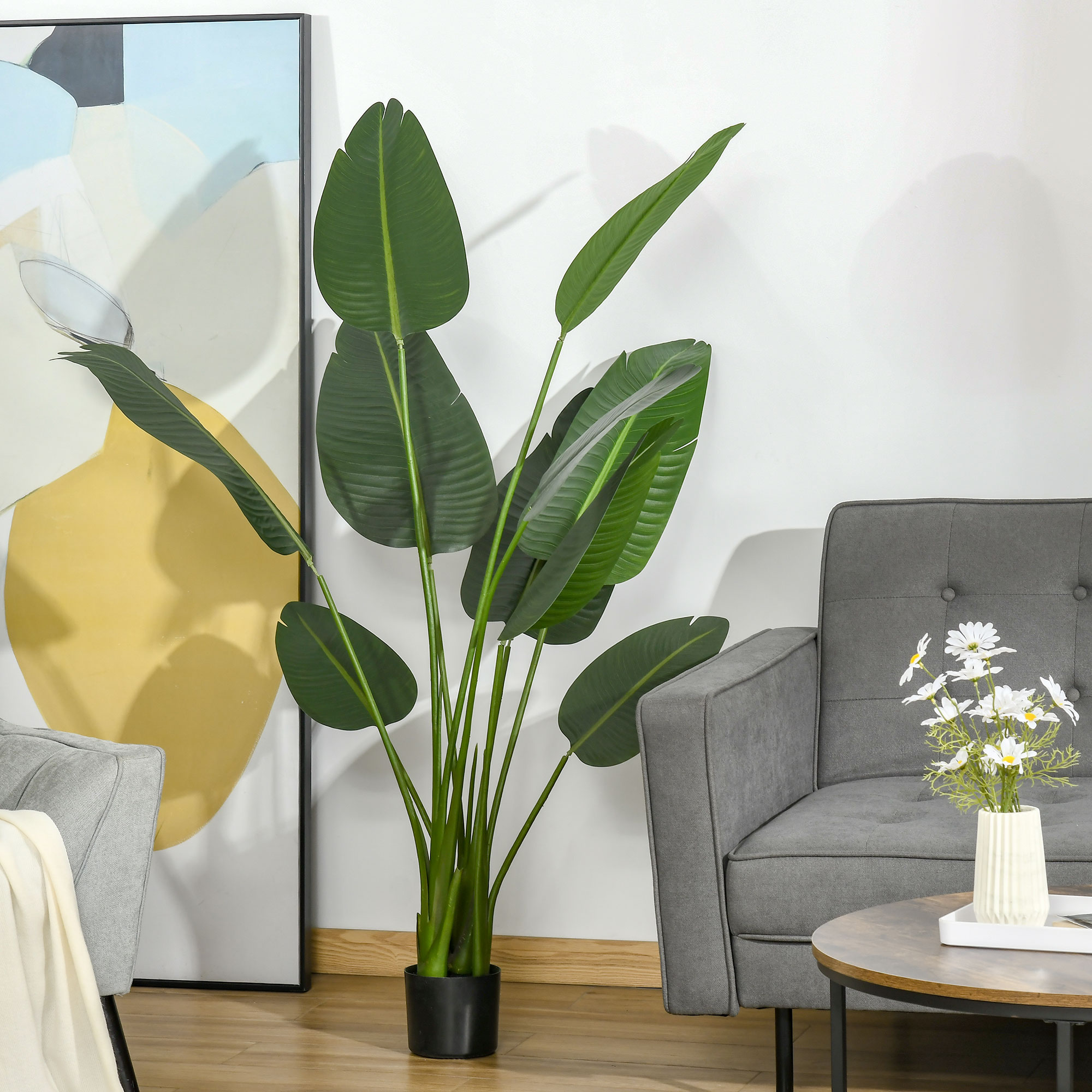 Una planta de plátano artificial alta en una maceta negra se encuentra entre un sofá gris, una obra de arte y una mesa pequeña.