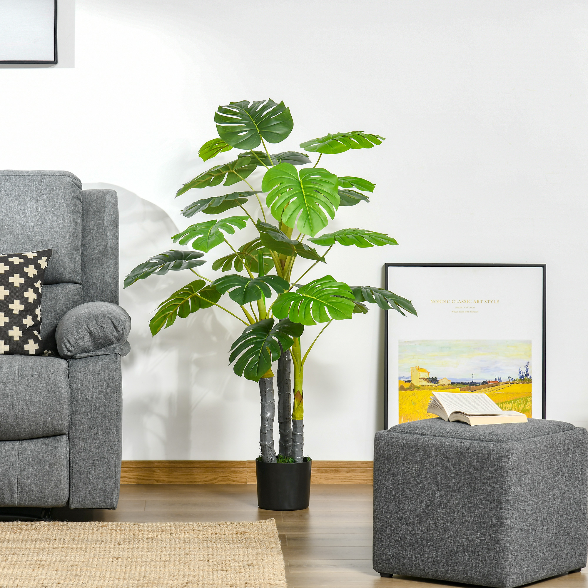 Una gran planta de monstera artificial en una maceta entre un sillón gris y un taburete a juego.