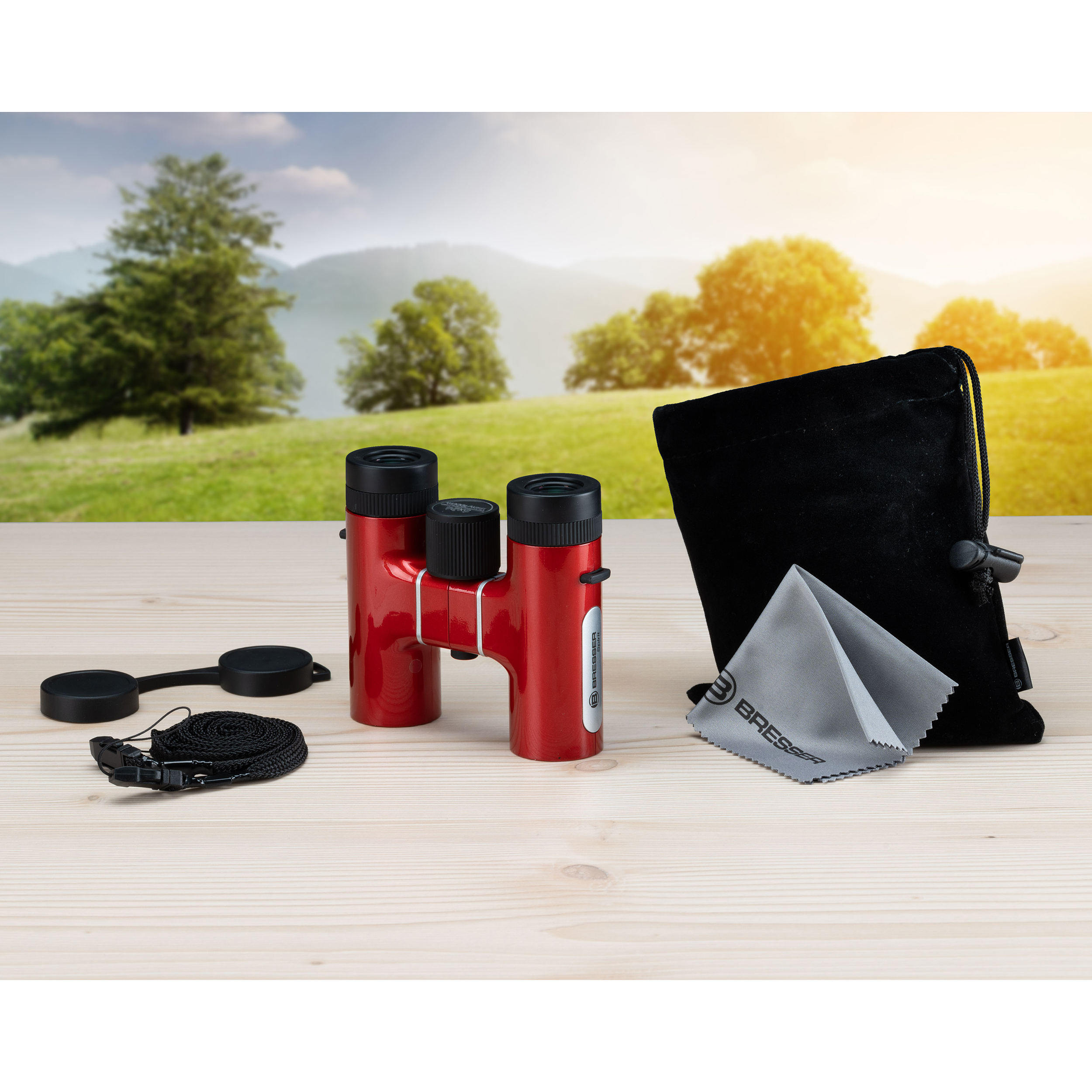 Binocolo rosso con accessori, custodia e panno su tavolo in legno.
