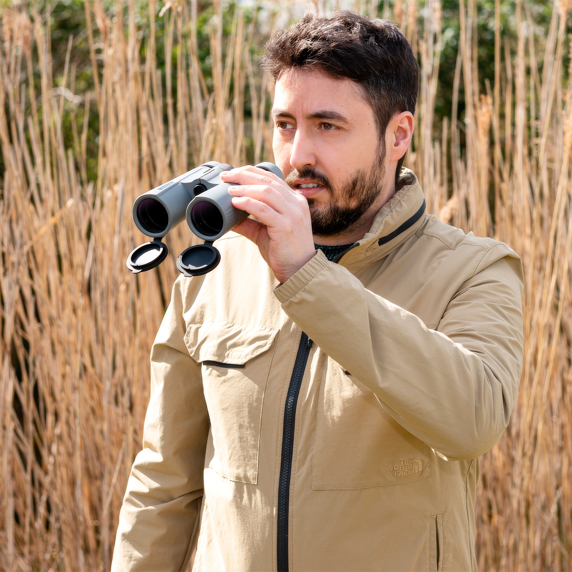 Un uomo barbuto con giacca beige tiene un binocolo all'aperto con erba alta sullo sfondo.
