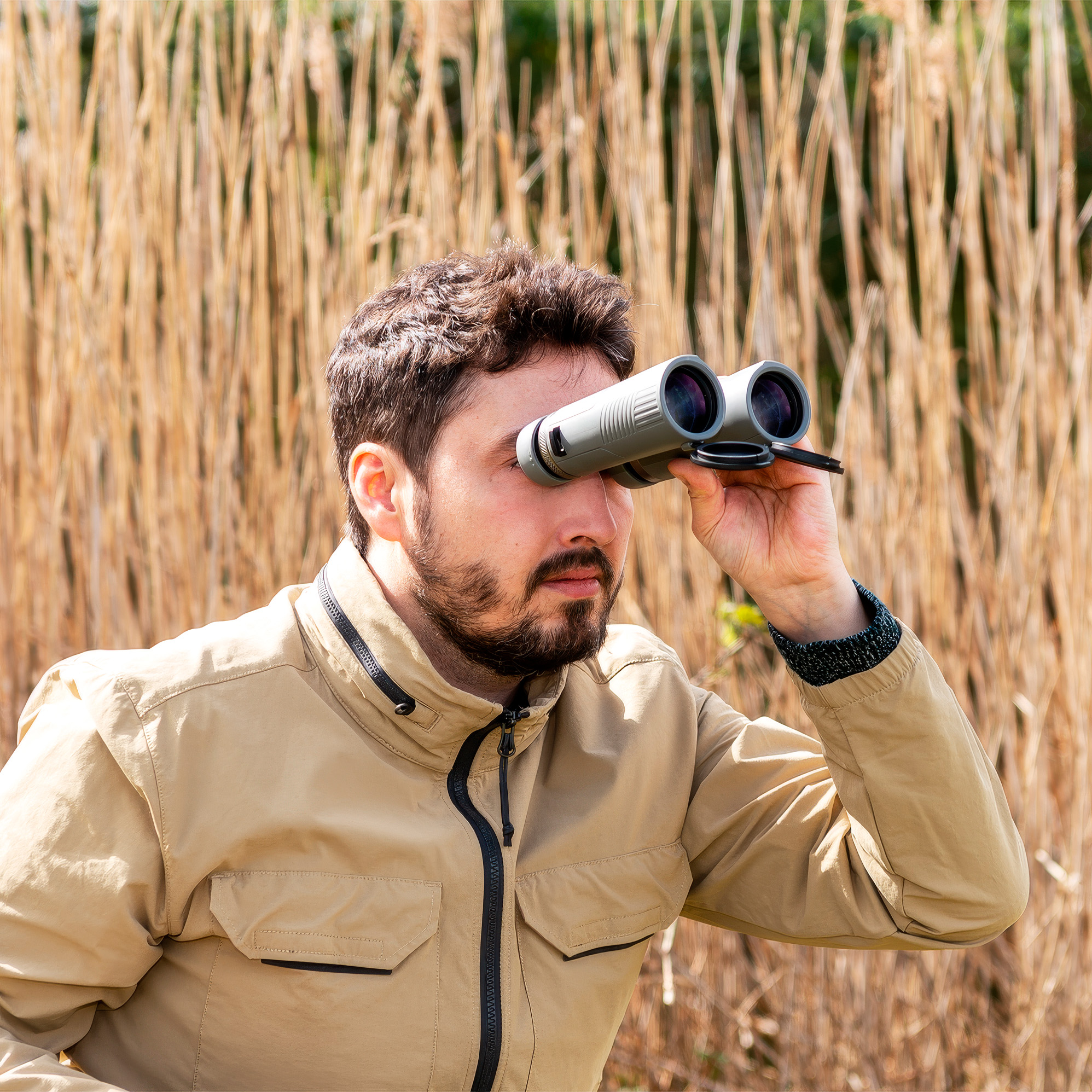 Un uomo barbuto con una giacca beige guarda attraverso un binocolo grigio su uno sfondo di canne secche.