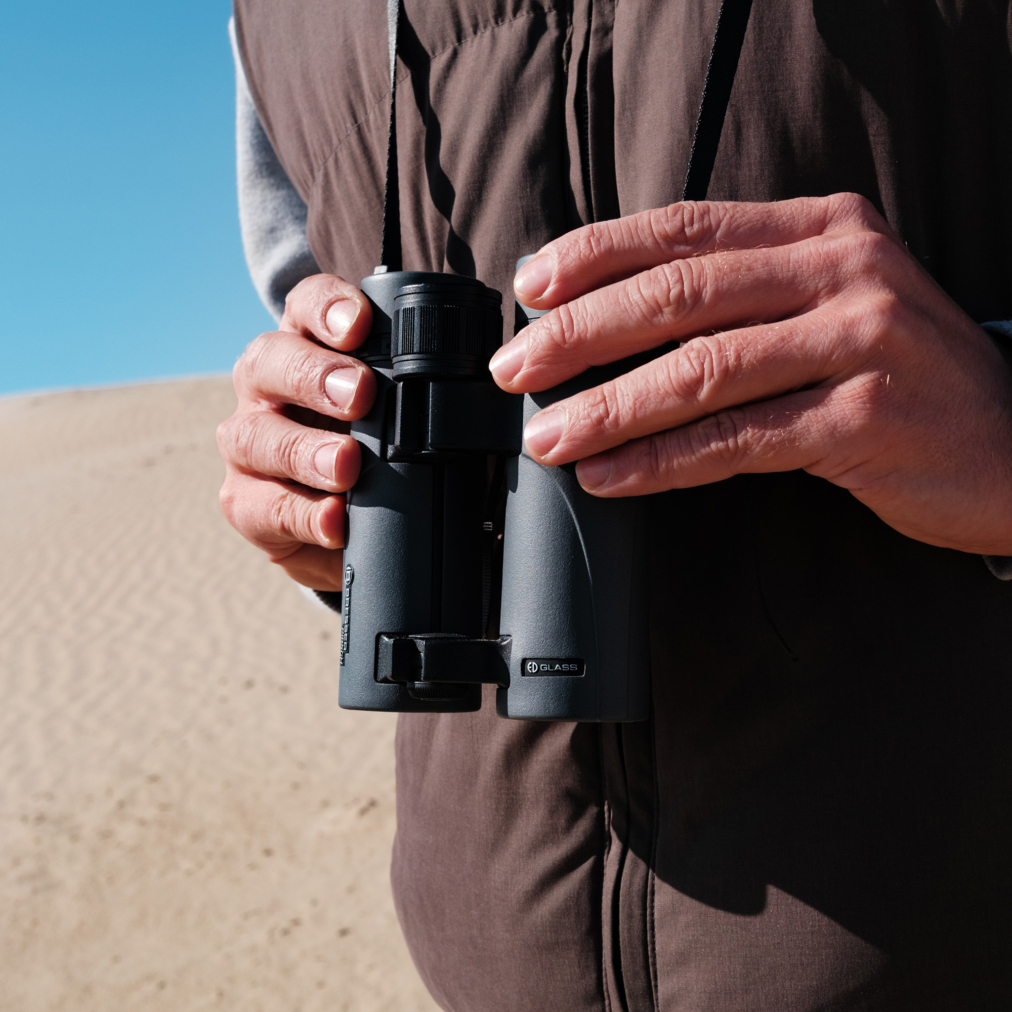 Una persona con chaleco marrón sostiene binoculares negros con fondo de desierto y cielo azul.