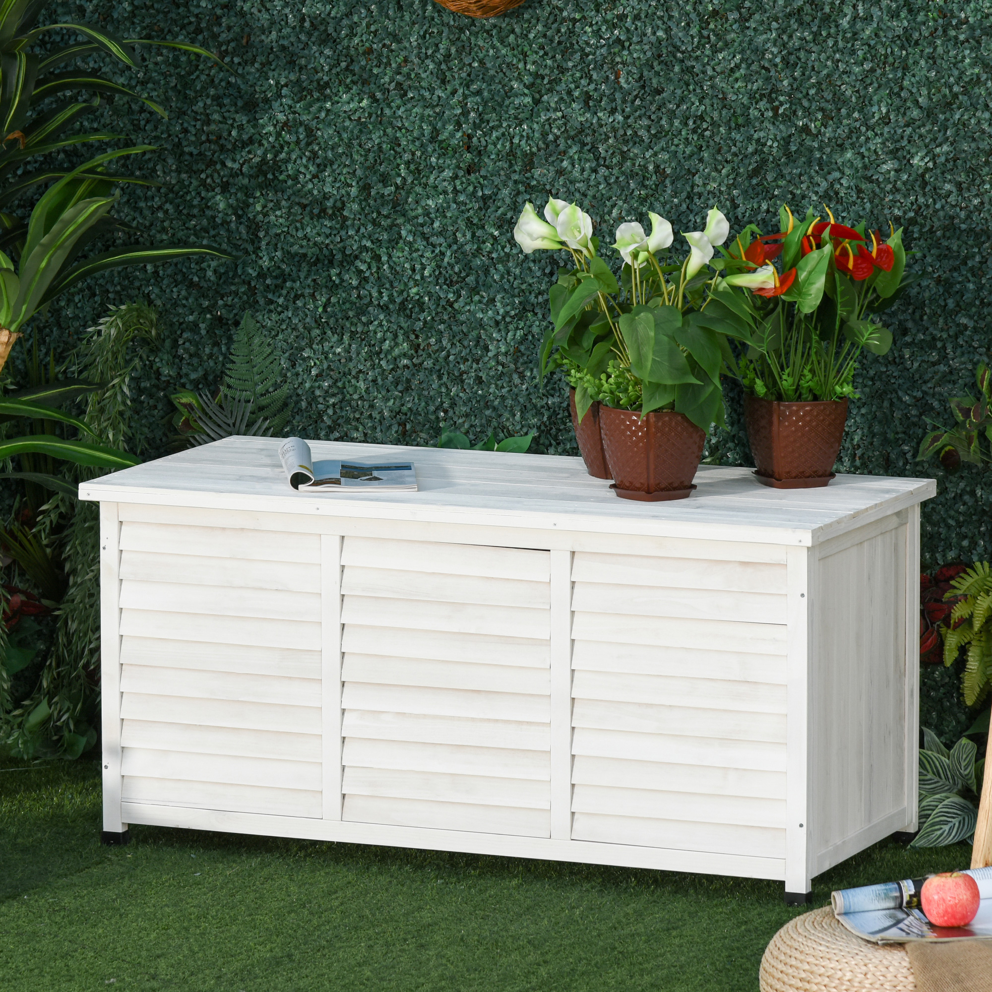 Un baúl de madera blanca sobre césped verde con macetas encima, frente a una pared verde artificial.