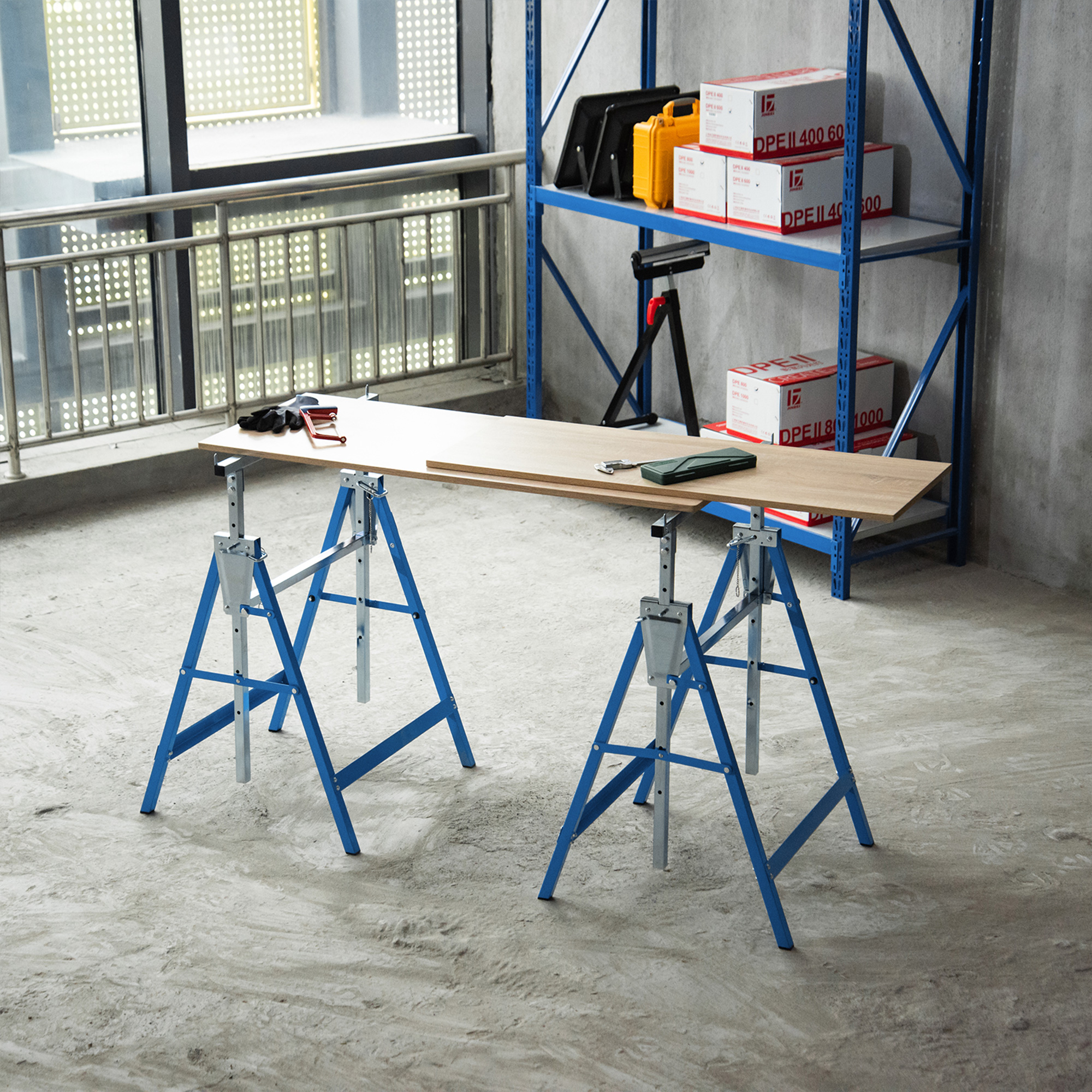 Dos caballetes azules sostienen un tablero de madera en un suelo de hormigón, con estantes y cajas al fondo.