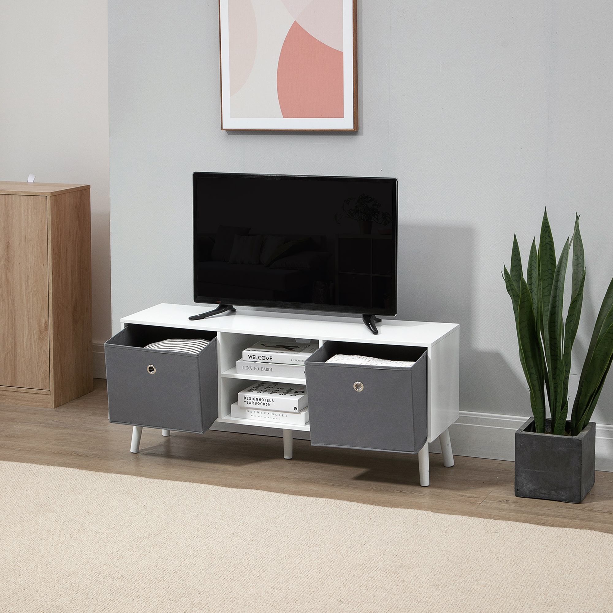 Un mueble de TV blanco con cajas de tela gris, un televisor encima, un cuadro enmarcado en la pared gris y una planta al lado.