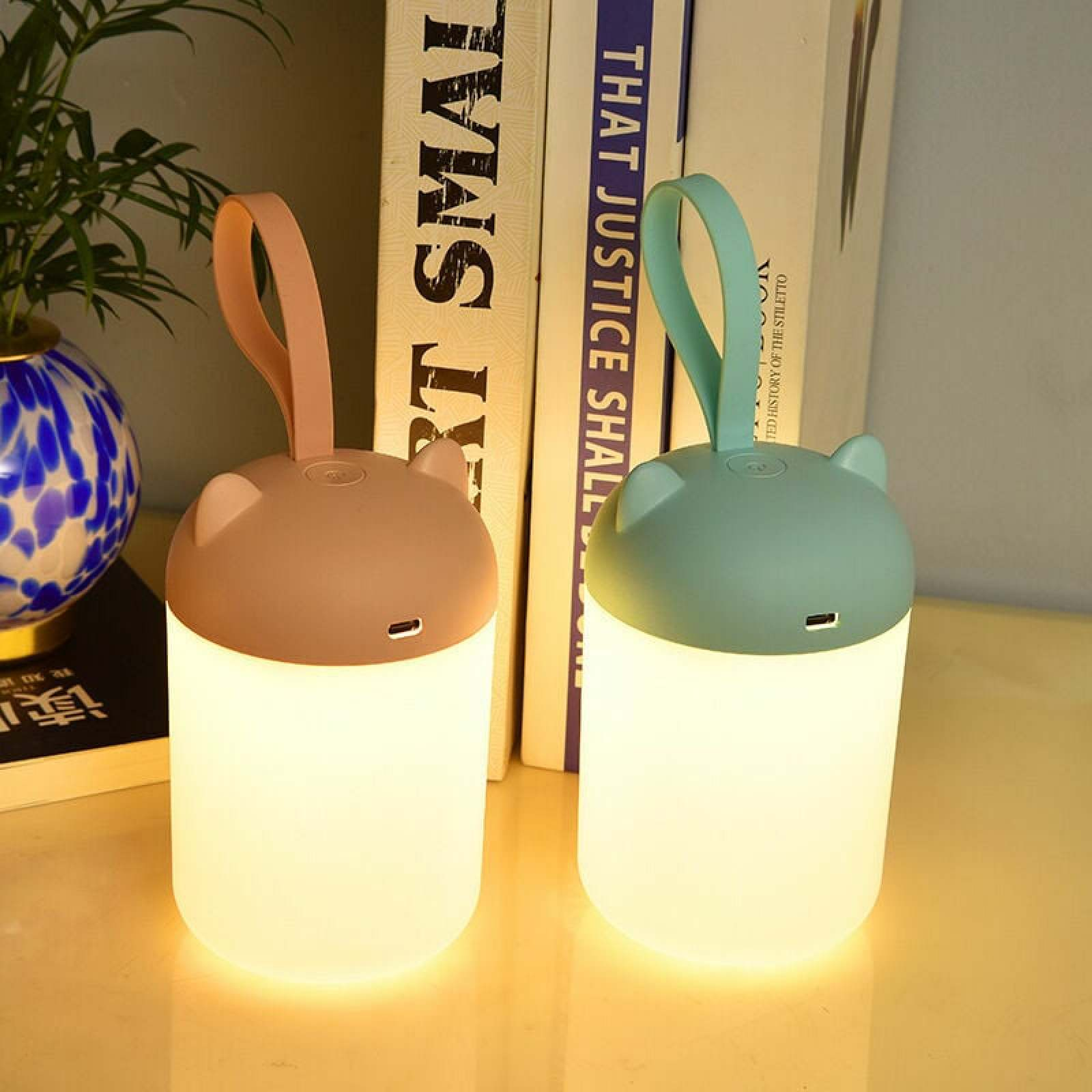 Zwei kleine, katzenohrige Lampen in Rosa und Türkis stehen auf einem Tisch vor Büchern.