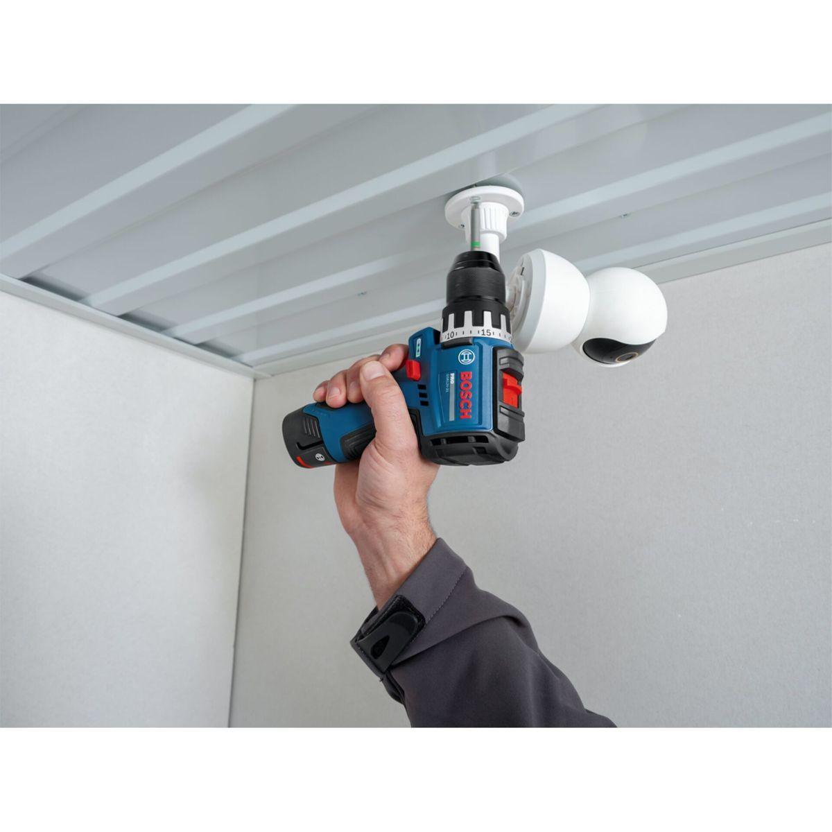 Una mano usa un trapano a batteria Bosch blu per montare una telecamera di sicurezza al soffitto.