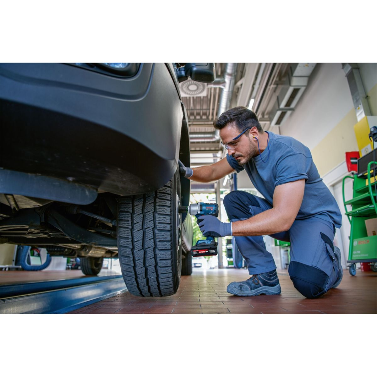 Un meccanico in abiti da lavoro grigi si inginocchia accanto a un'auto, usando un attrezzo sulla ruota.