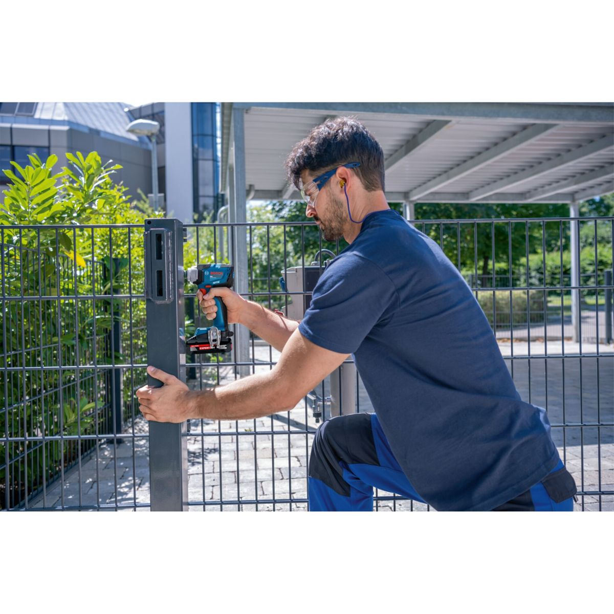 Un uomo in abiti da lavoro blu usa un trapano a batteria su un palo di recinzione.