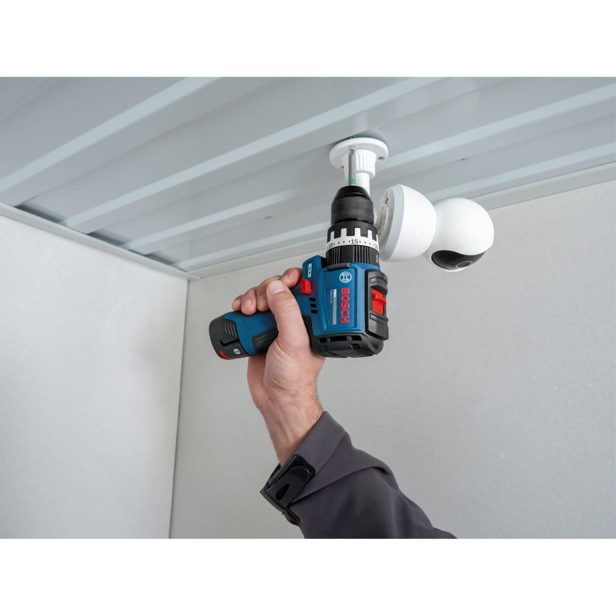 Una persona usa un trapano Bosch blu per montare una telecamera di sicurezza bianca su un soffitto.