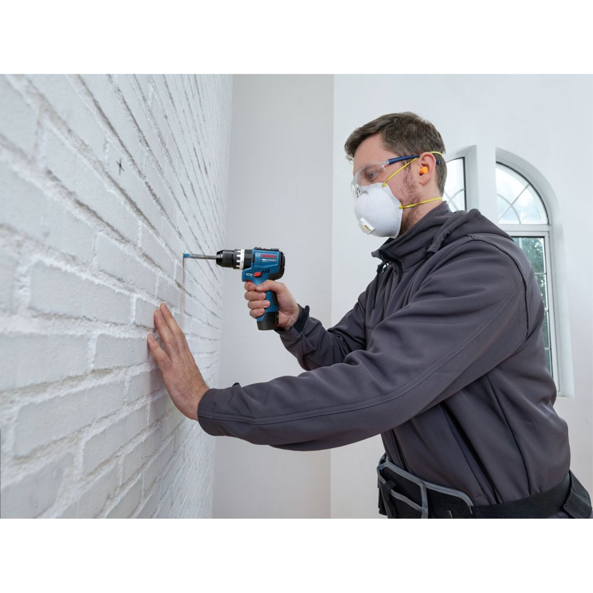 Un lavoratore con occhiali di protezione e mascherina fora un muro di mattoni bianchi.