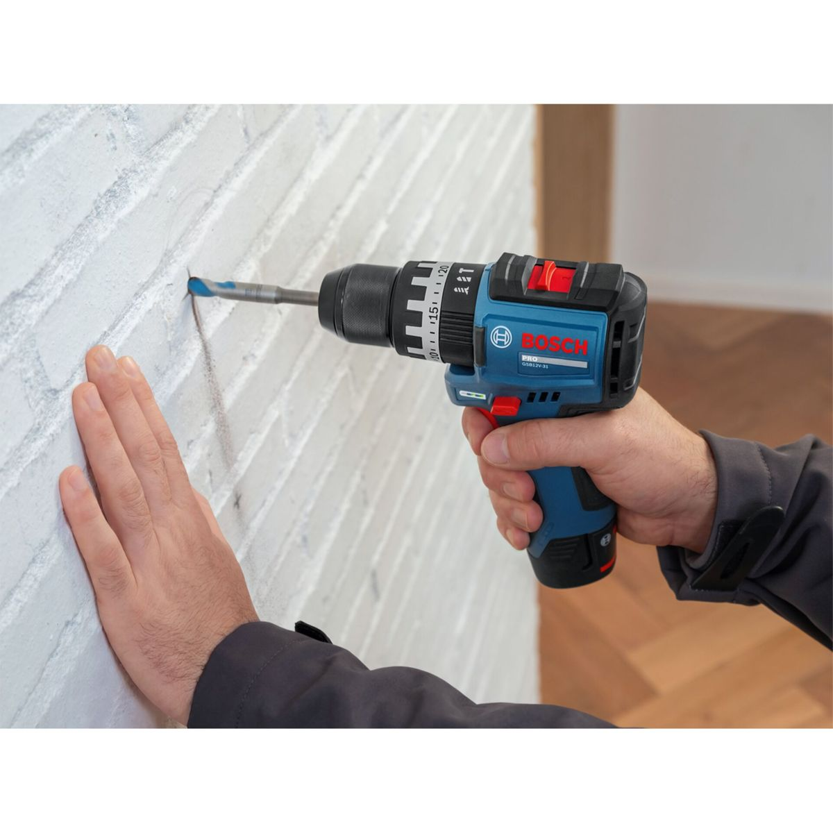 Una mano tiene un trapano a batteria Bosch blu che fora un muro di mattoni bianchi.