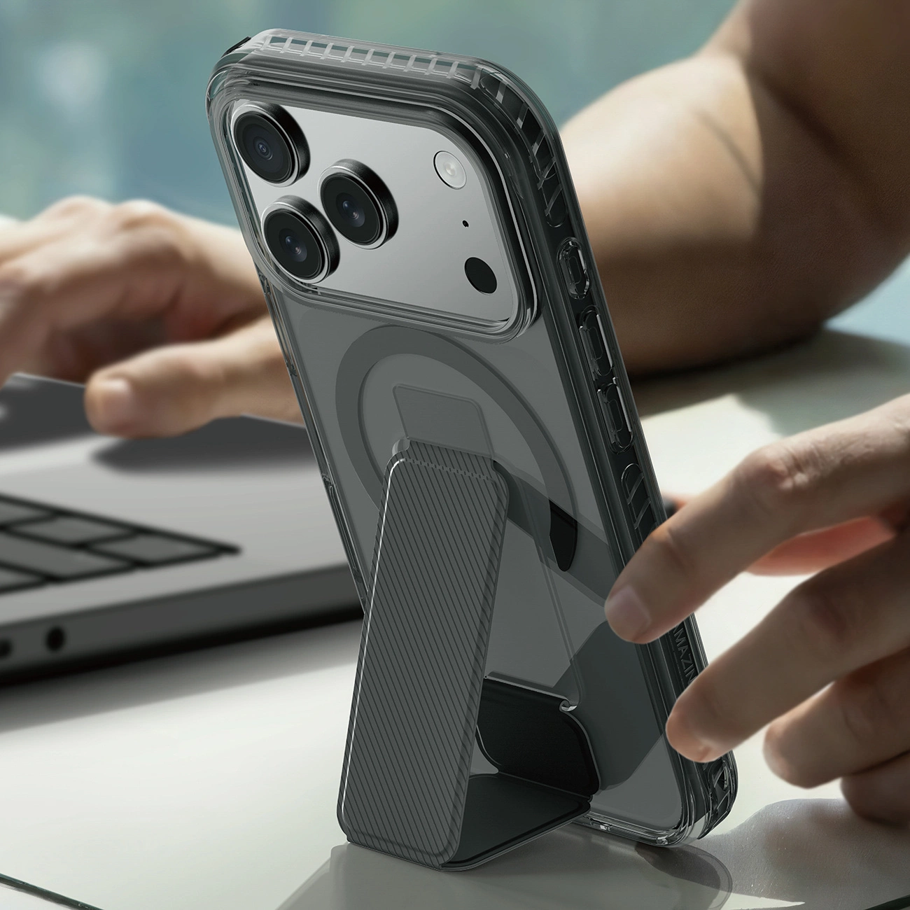 Ein Smartphone in einer Klarsichthülle mit integriertem Ständer steht aufrecht auf einem Schreibtisch neben einem Laptop.