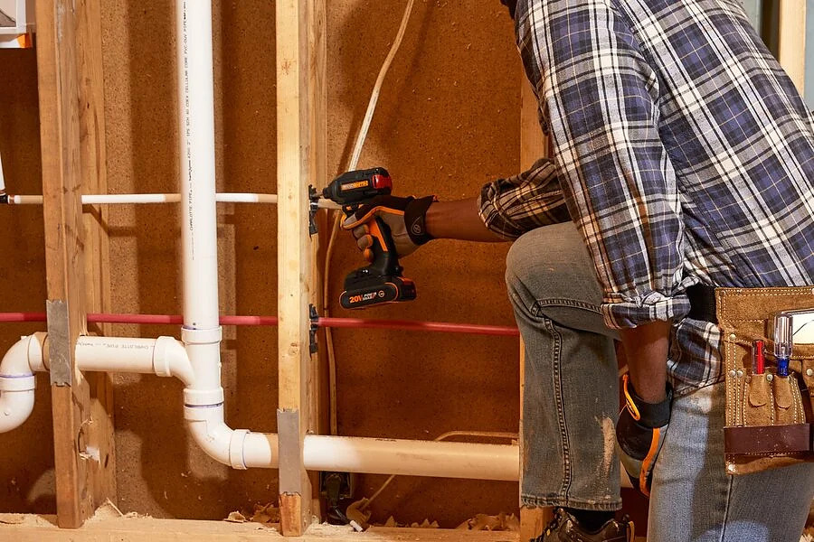 Un lavoratore in camicia a quadri usa un trapano su montanti in legno vicino ai tubi.