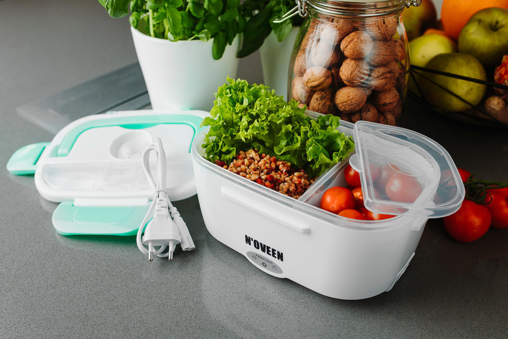 Weiße elektrische Lunchbox mit Salat und Tomaten, umgeben von Pflanzen, Walnüssen im Glas und frischem Obst.