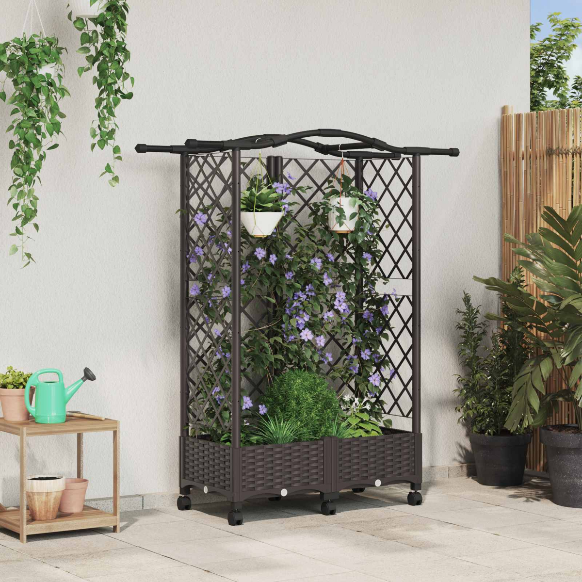 Ein schwarzes Spalier mit Rädern, bewachsen mit violetten Blumen, steht auf einer Terrasse neben Pflanzen und einer Gießkanne.