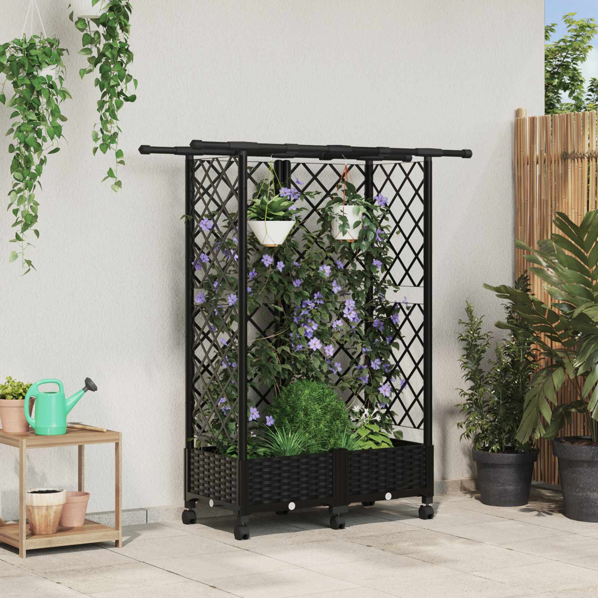 Ein schwarzes Metallgitter mit lila blühenden Ranken auf Rädern, auf einer Terrasse neben Pflanzen und einem Hocker.
