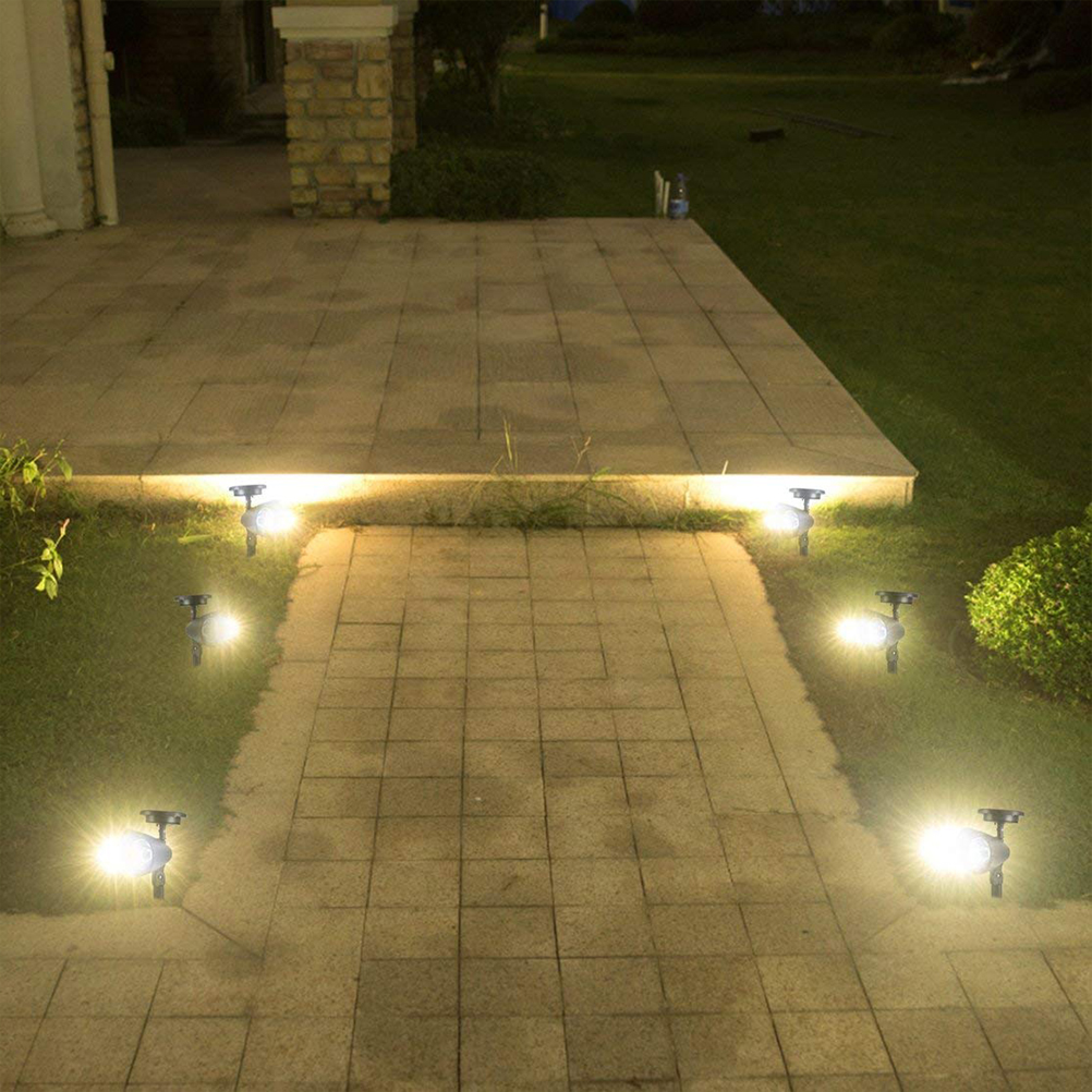 Sechs solarbetriebene Spotlights beleuchten abends einen gepflasterten Gartenweg und eine Terrasse.