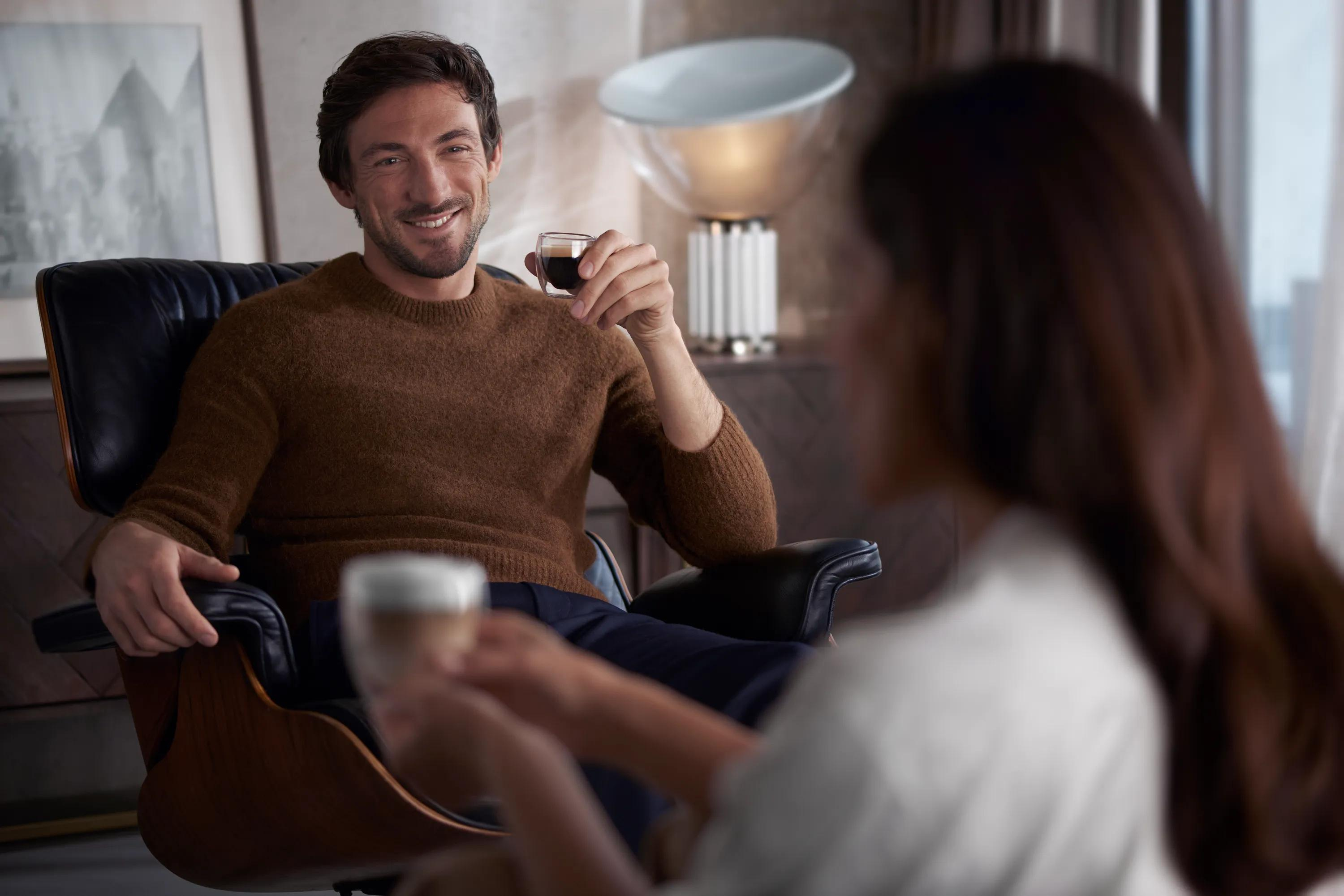 Uomo con maglione marrone siede su poltrona nera tenendo tazza di caffè mentre guarda una donna.