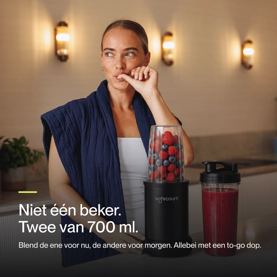 Een vrouw in blauwe jas staat naast een blender vol bessen en een smoothiebeker.