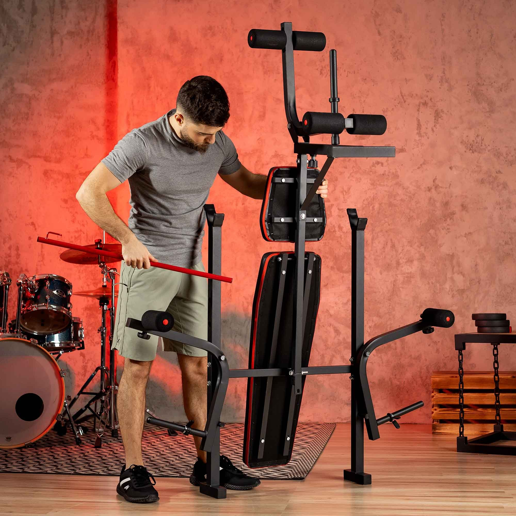 Ein Mann im grauen T-Shirt baut ein multifunktionales Fitnessgerät in einem Raum mit Drumset auf.