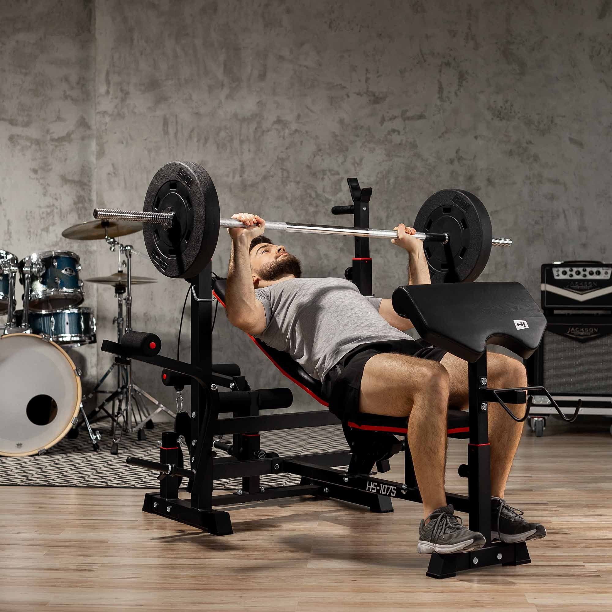 Ein Mann im Fitnessstudio drückt eine Hantel auf einer Schrägbank, mit Schlagzeug und Verstärker im Hintergrund.