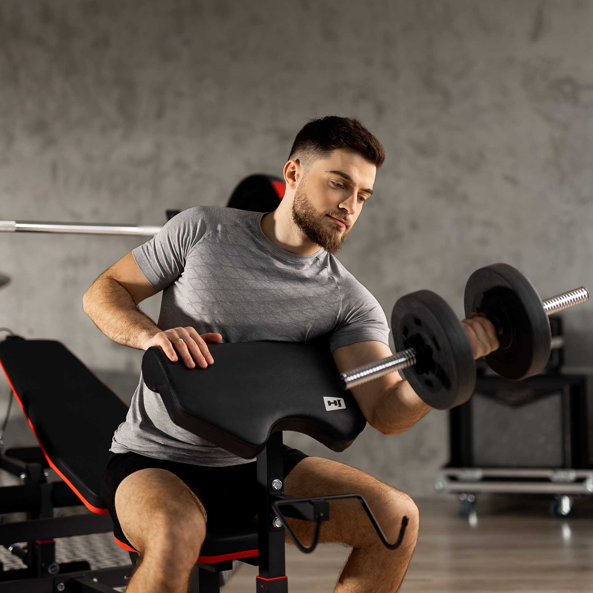 Ein Mann mit Bart sitzt auf einer Fitnessbank und hebt eine Hantel in einem Raum mit grauer Wand.