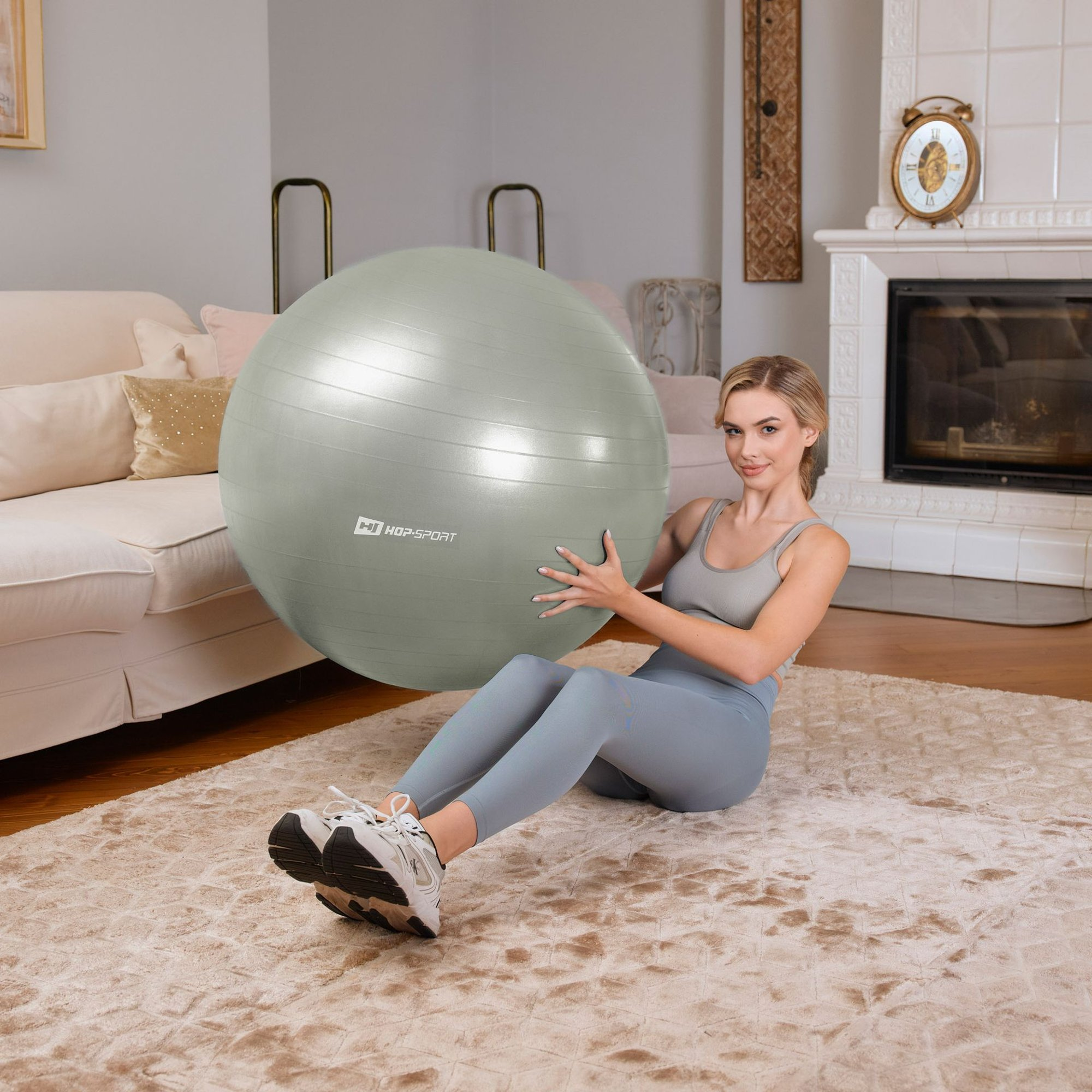 Eine blonde Frau in Sportkleidung sitzt auf einem Teppich in einem Wohnzimmer und hält einen großen grauen Gymnastikball.