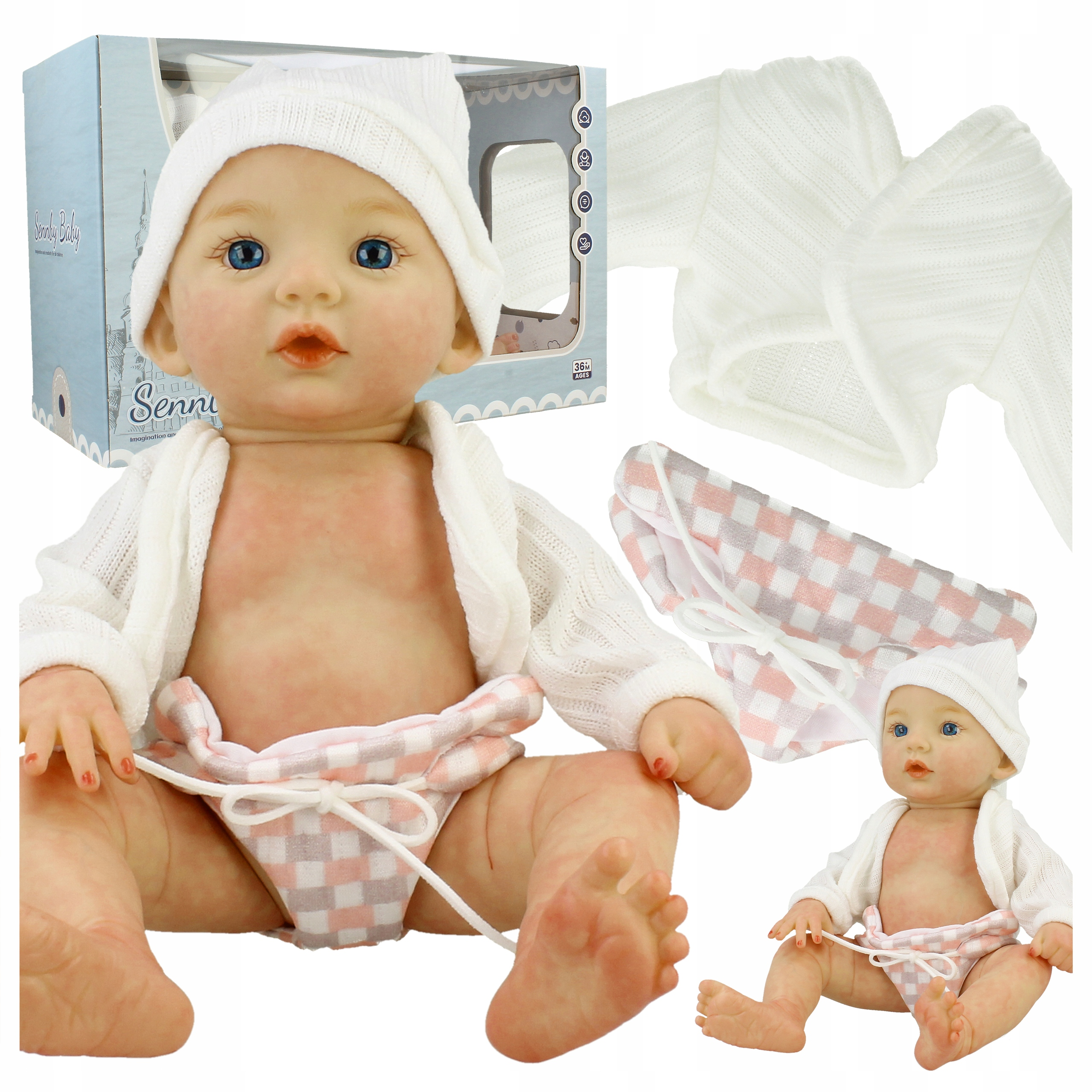 Lalka reborn z niebieskimi oczami, białą czapką, kardiganem, w kratkę pieluszką, pudełkiem i ubrankami.