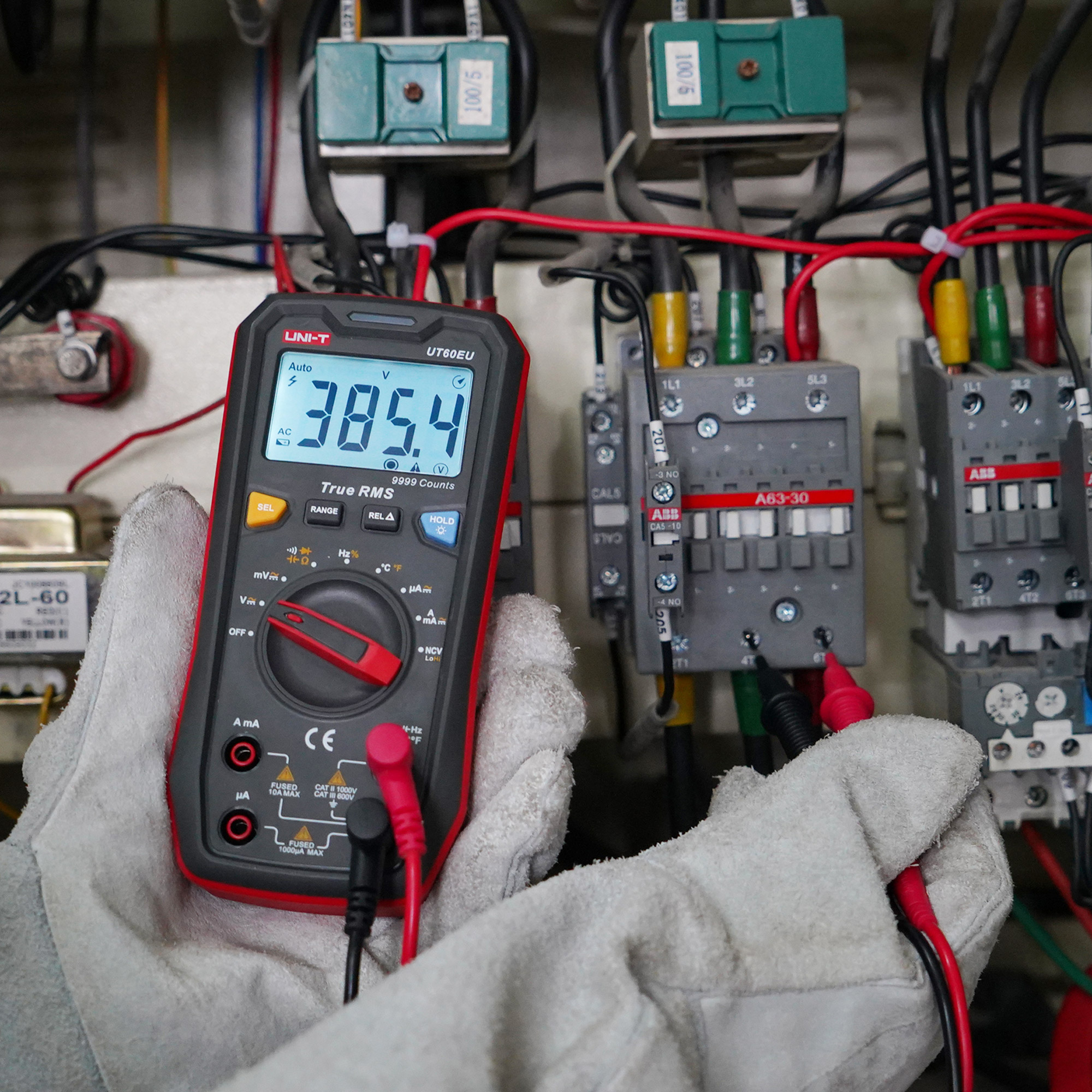 Ręka w rękawici trzyma multimetr wskazujący 385,4 V przed panelem elektrycznym.