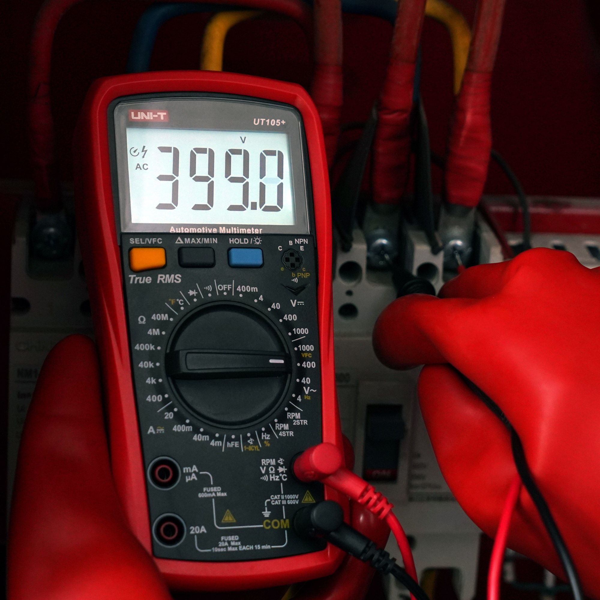 W rękawiczce dłoń trzyma czerwony multimetr wskazujący 399,0V przed kablami elektrycznymi.