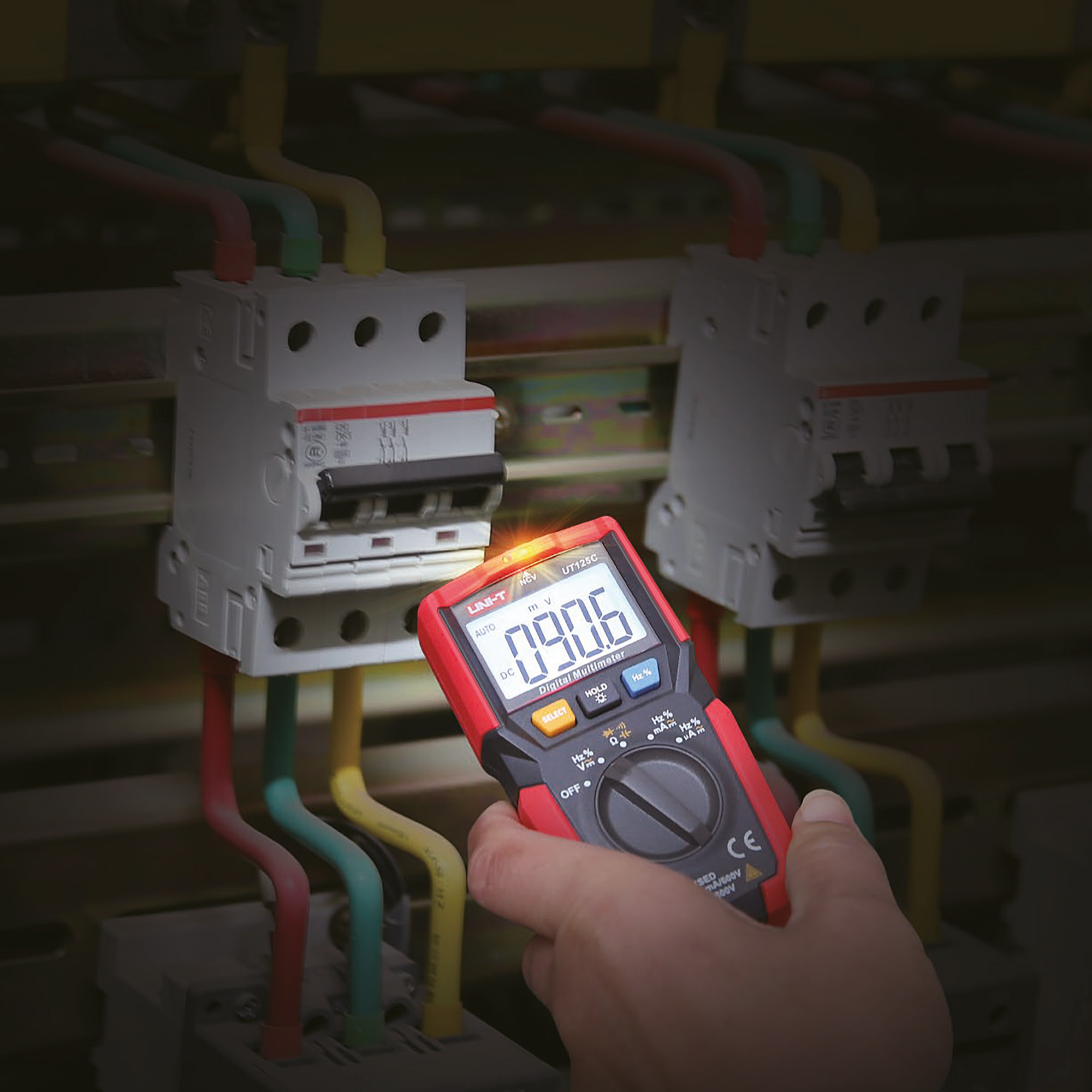 Ręka trzyma czerwony multimetr cyfrowy mierzący wyłączniki elektryczne z kolorowymi przewodami.