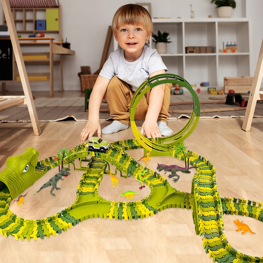 Mały chłopiec klęczy na podłodze, uśmiechając się, bawiąc się zielonym torem wyścigowym z dinozaurami.