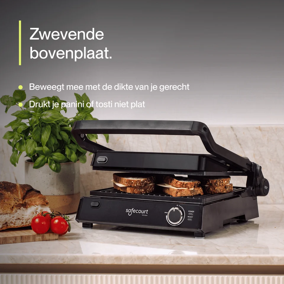 Gril de contact noir sur un plan de travail avec des sandwichs, une plante en pot et des tomates.