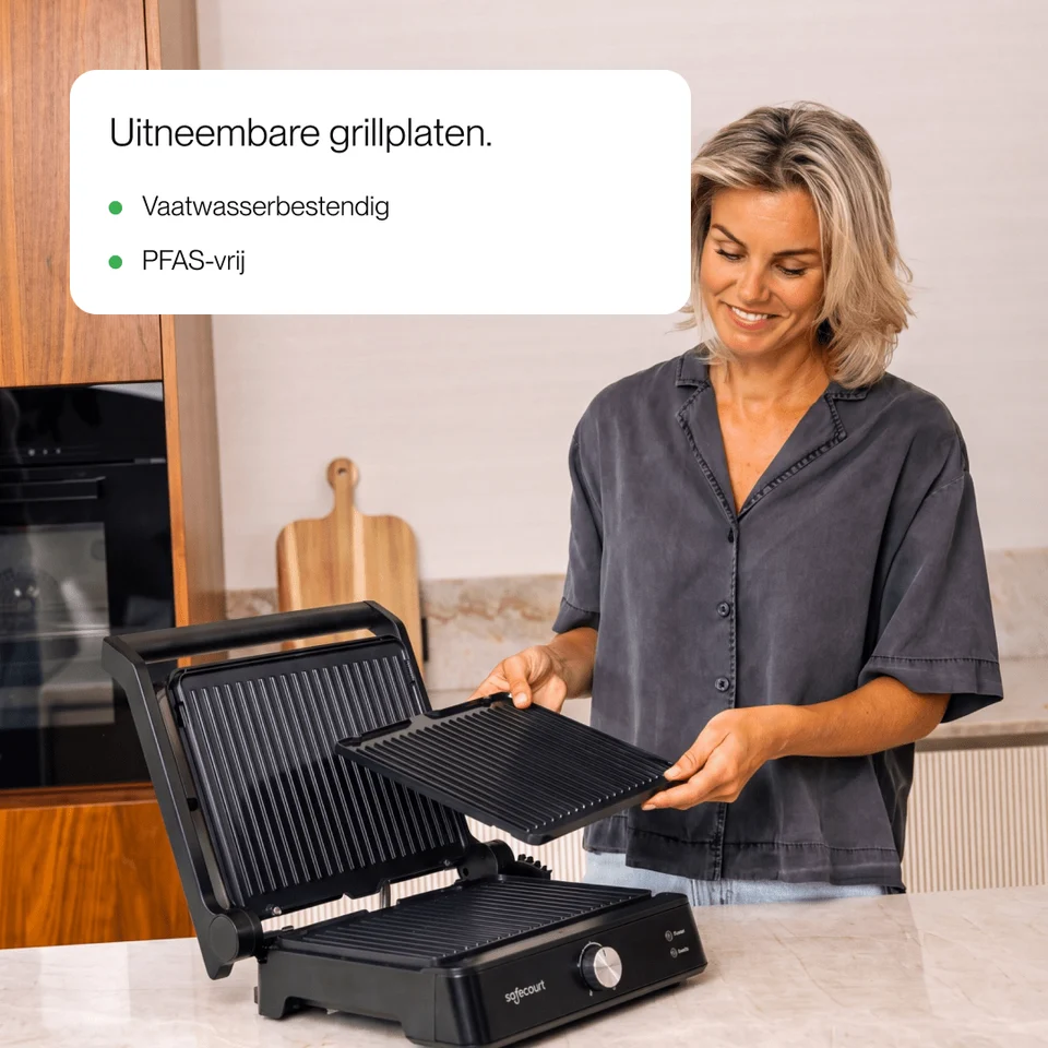 A smiling woman in a kitchen holds a removable grill plate from a black electric contact grill.