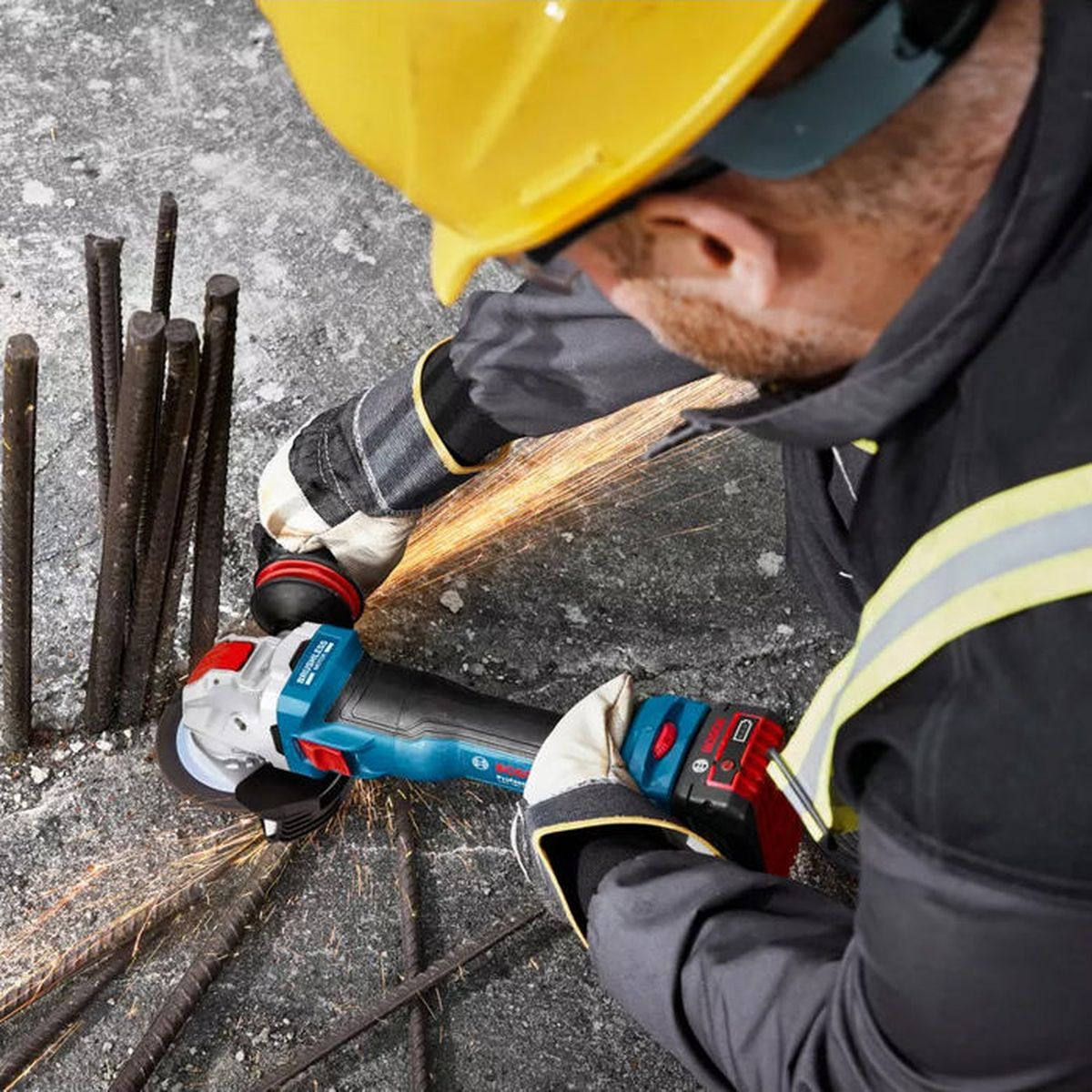 Ein Arbeiter in gelbem Helm schneidet Metallbewehrung mit einem Winkelschleifer, wodurch Funken sprühen.
