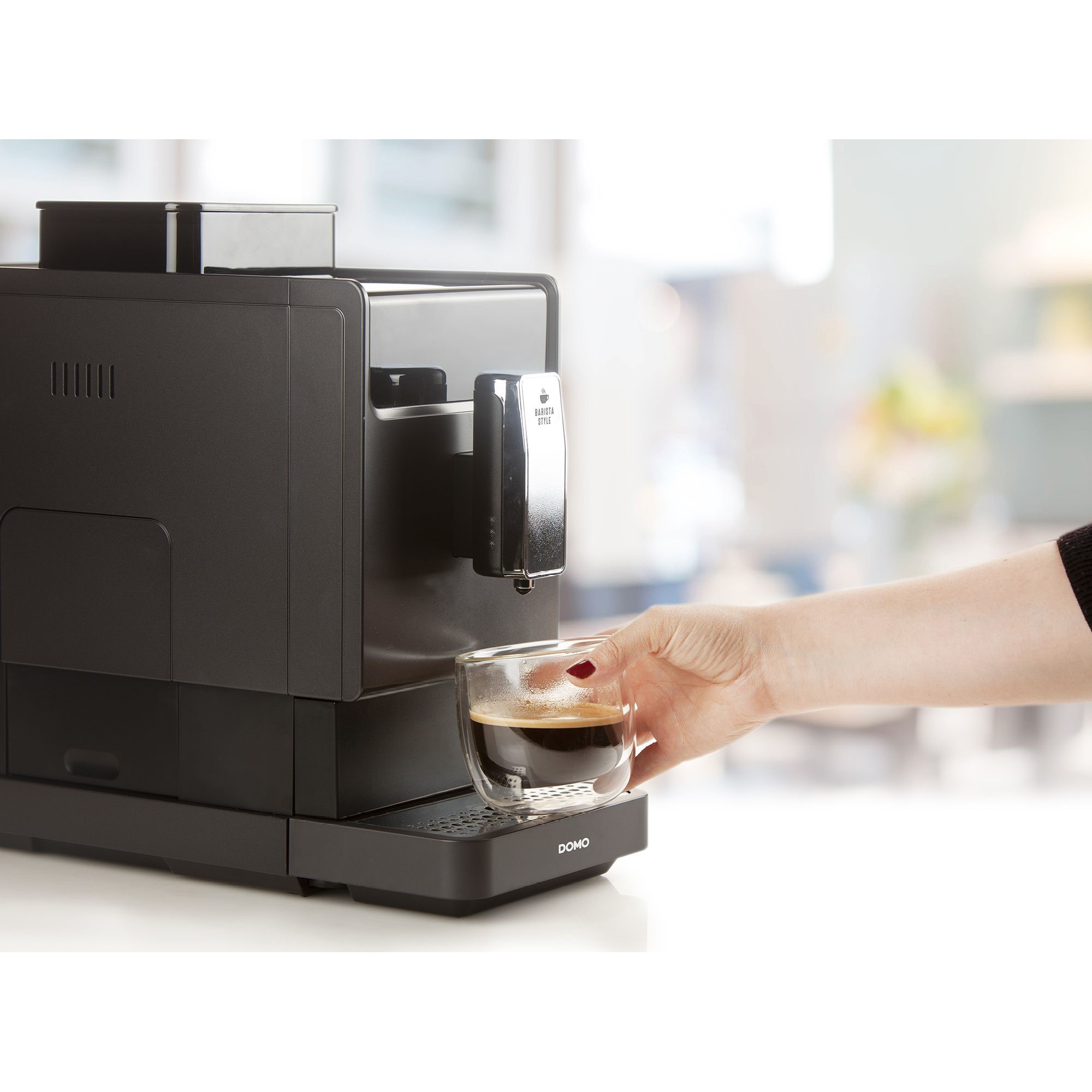 Una mano tiene una tazza di vetro di caffè appena fatto sotto una moderna macchina da caffè nera.