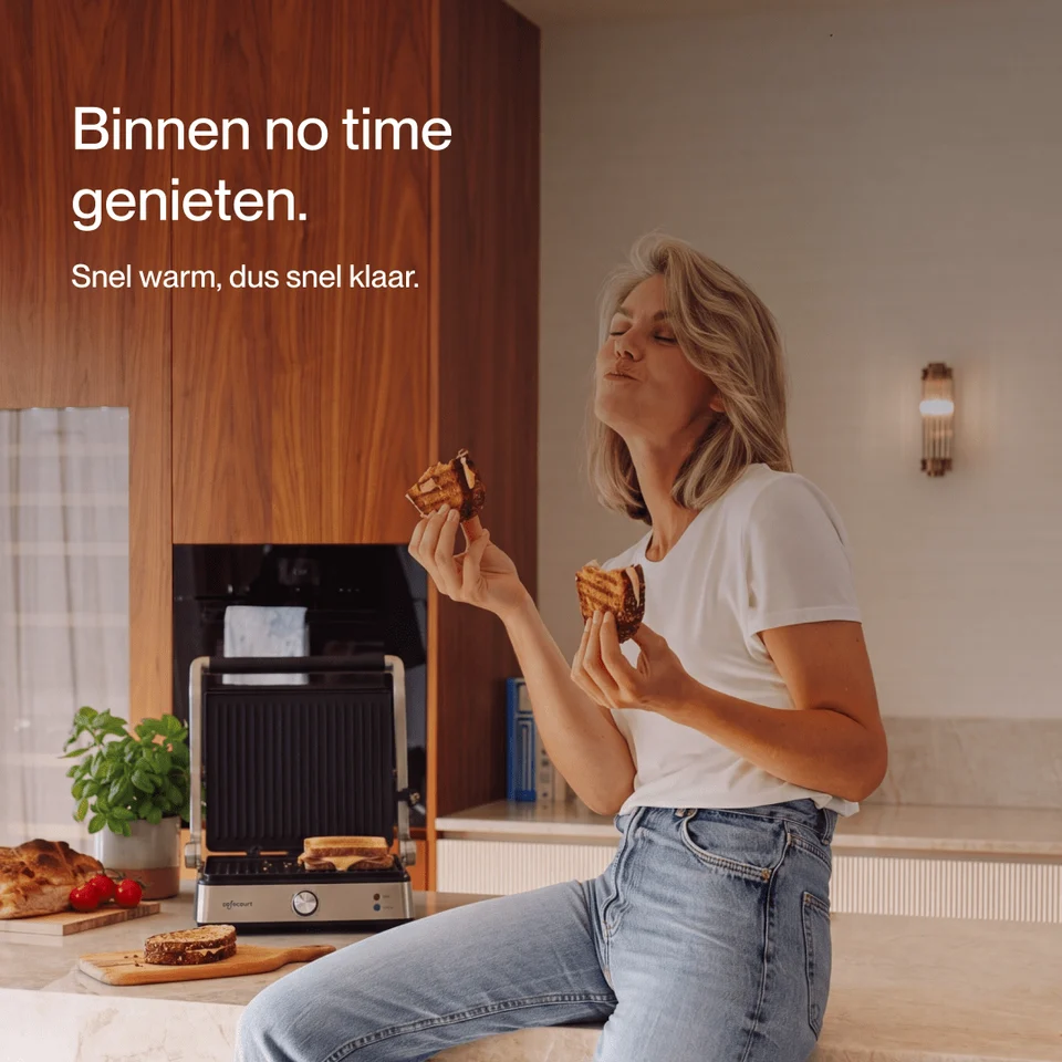 A blonde woman sits in a modern kitchen, happily eating a grilled cheese sandwich near a panini press.