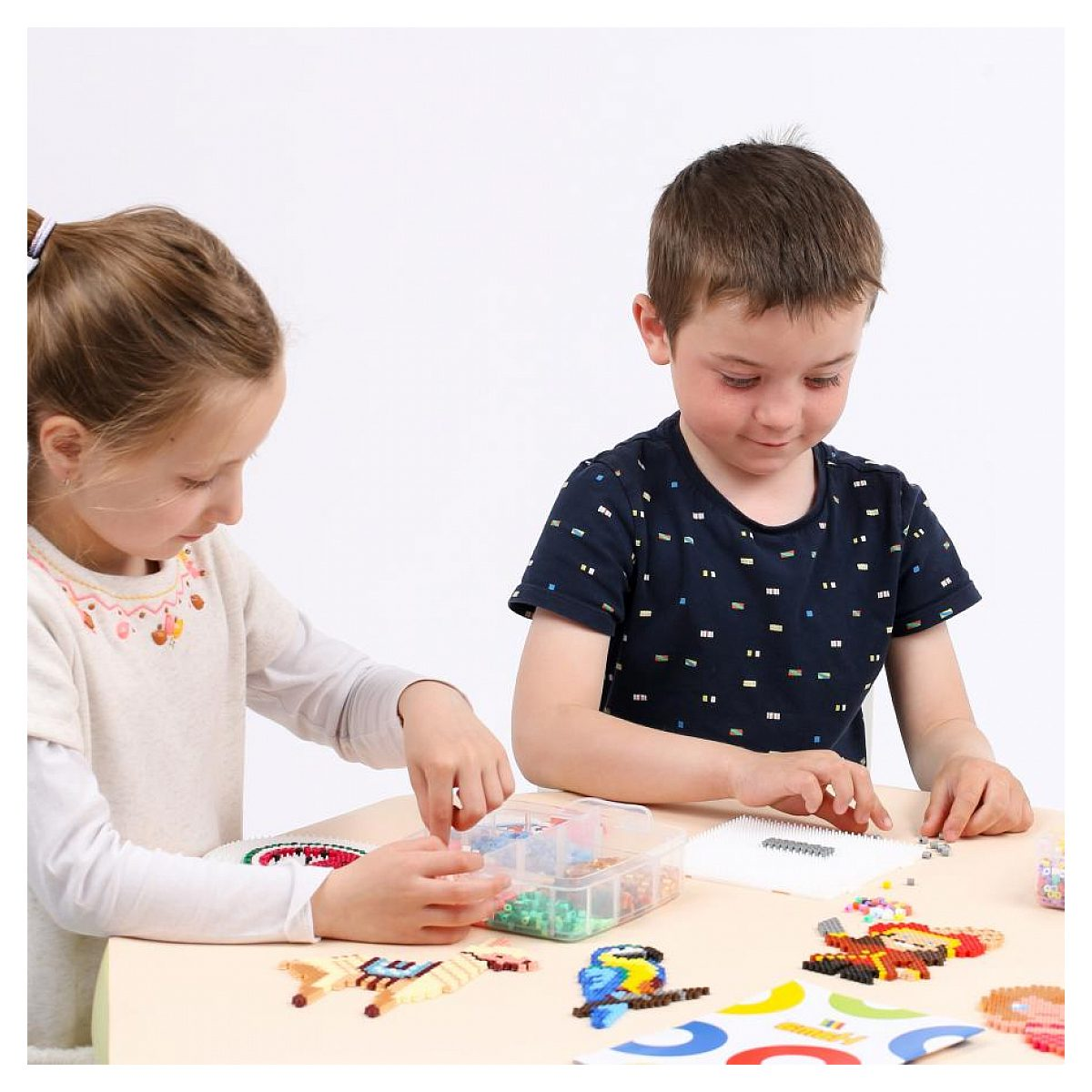 Ein junges Mädchen und ein Junge sitzen an einem Tisch, spielen mit bunten Steckperlen und erstellen verschiedene Muster.