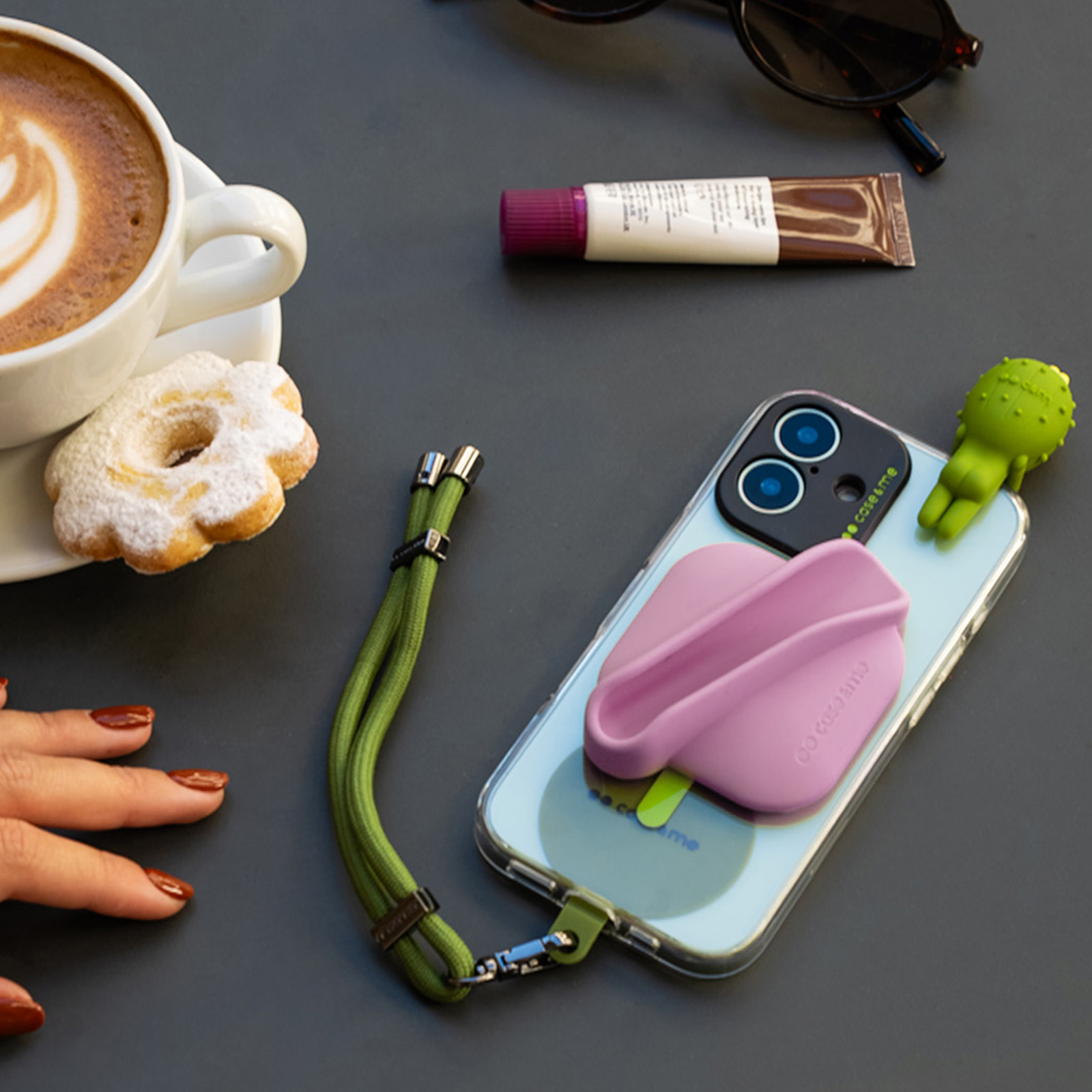 Un smartphone con correa verde, una taza de café con pastelito, gafas de sol y un tubo de crema en una mesa oscura.