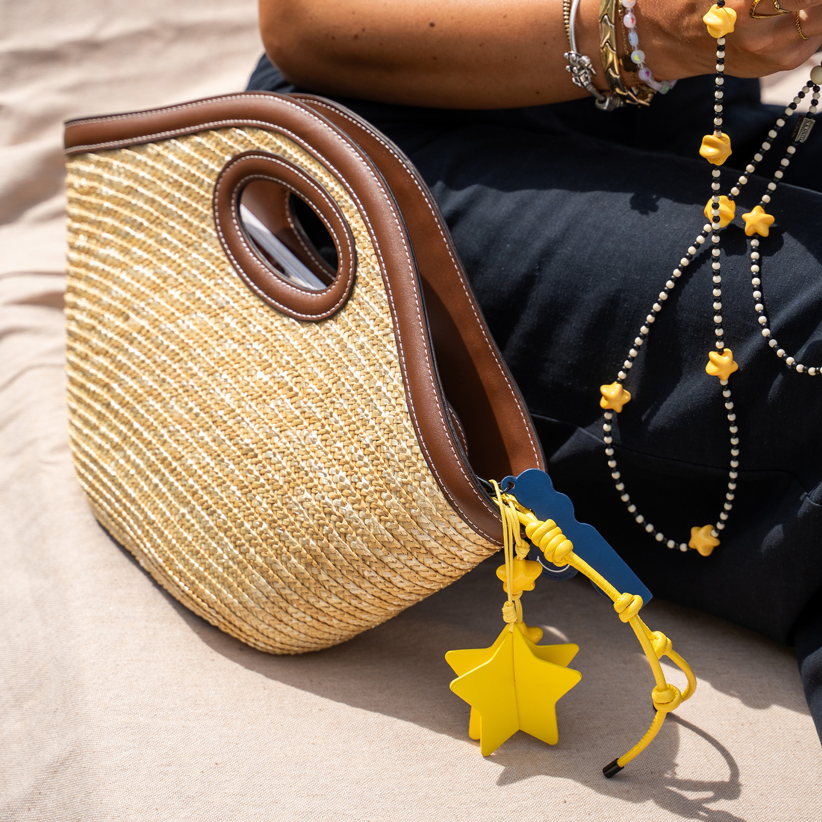 Bolso de paja con cuero marrón, amuleto de estrella amarilla y collar de cuentas sobre arena.
